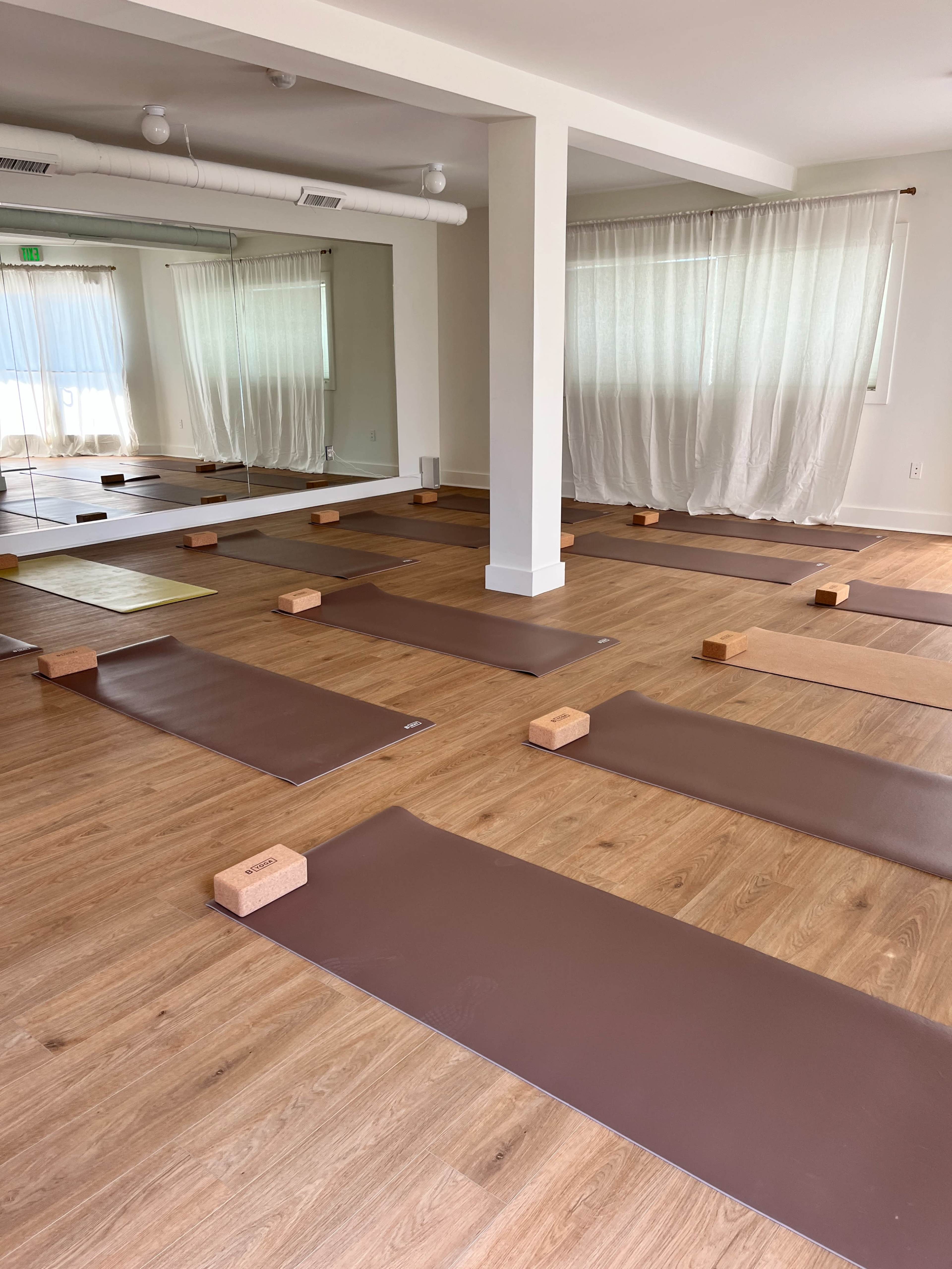 A yoga studio with several yoga mats arranged on a wooden floor, each with a wooden block placed beside them, and large windows covered by sheer curtains.