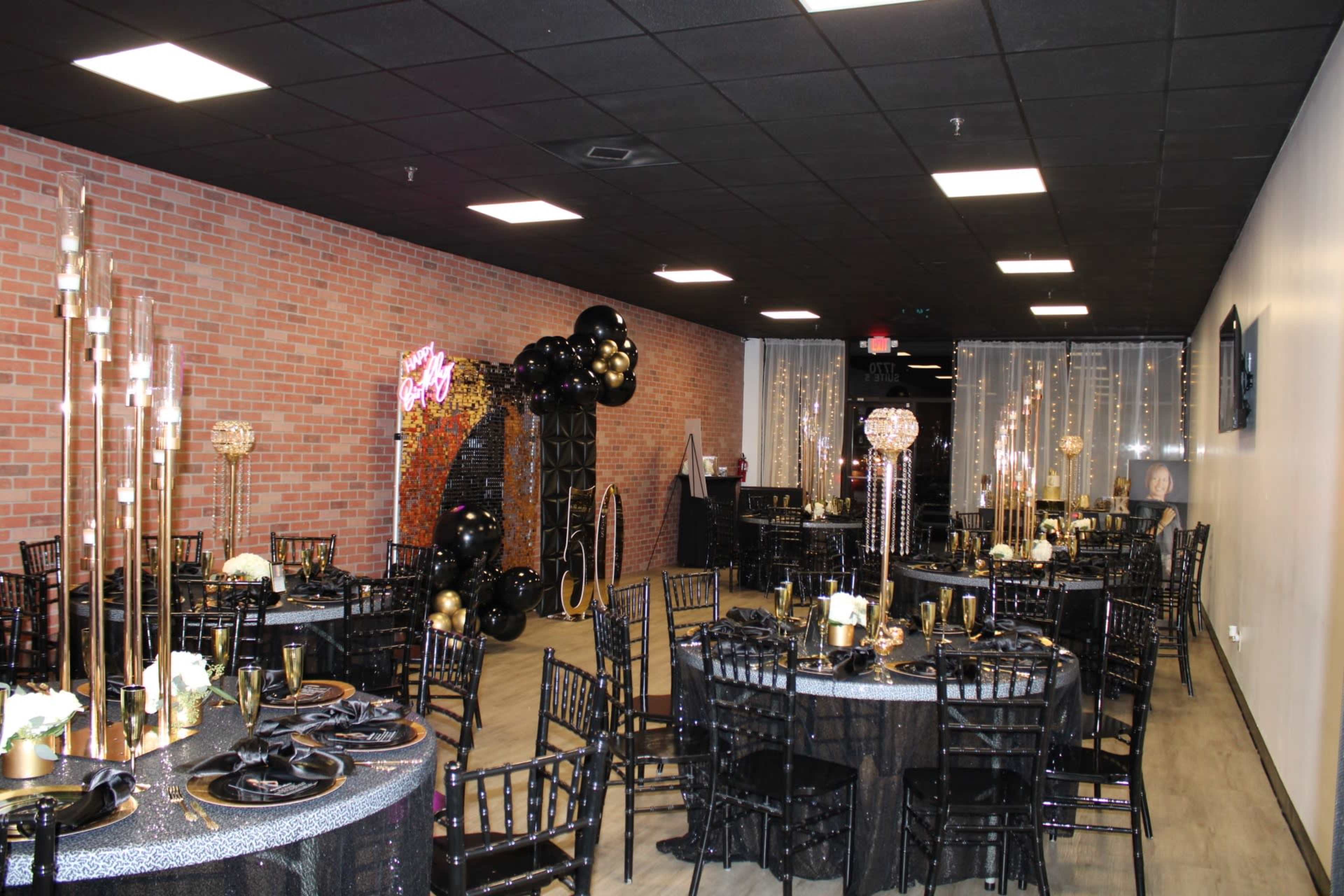 A decorated event space featuring black and gold themed tables, chairs, and decor, with a backdrop of balloons and lights.