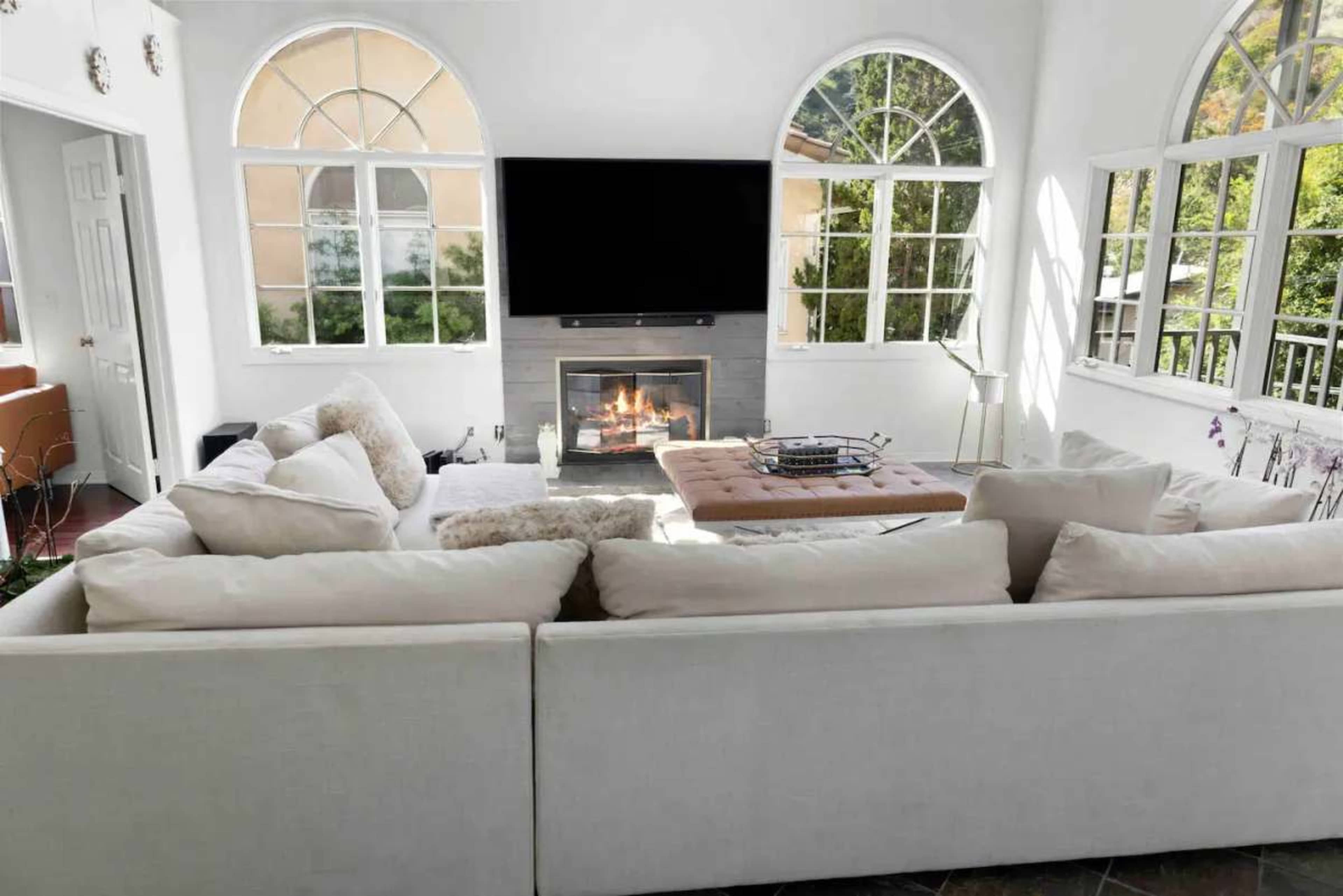 A spacious living room features a large sectional sofa facing a fireplace below a wall-mounted TV, framed by large arched windows.