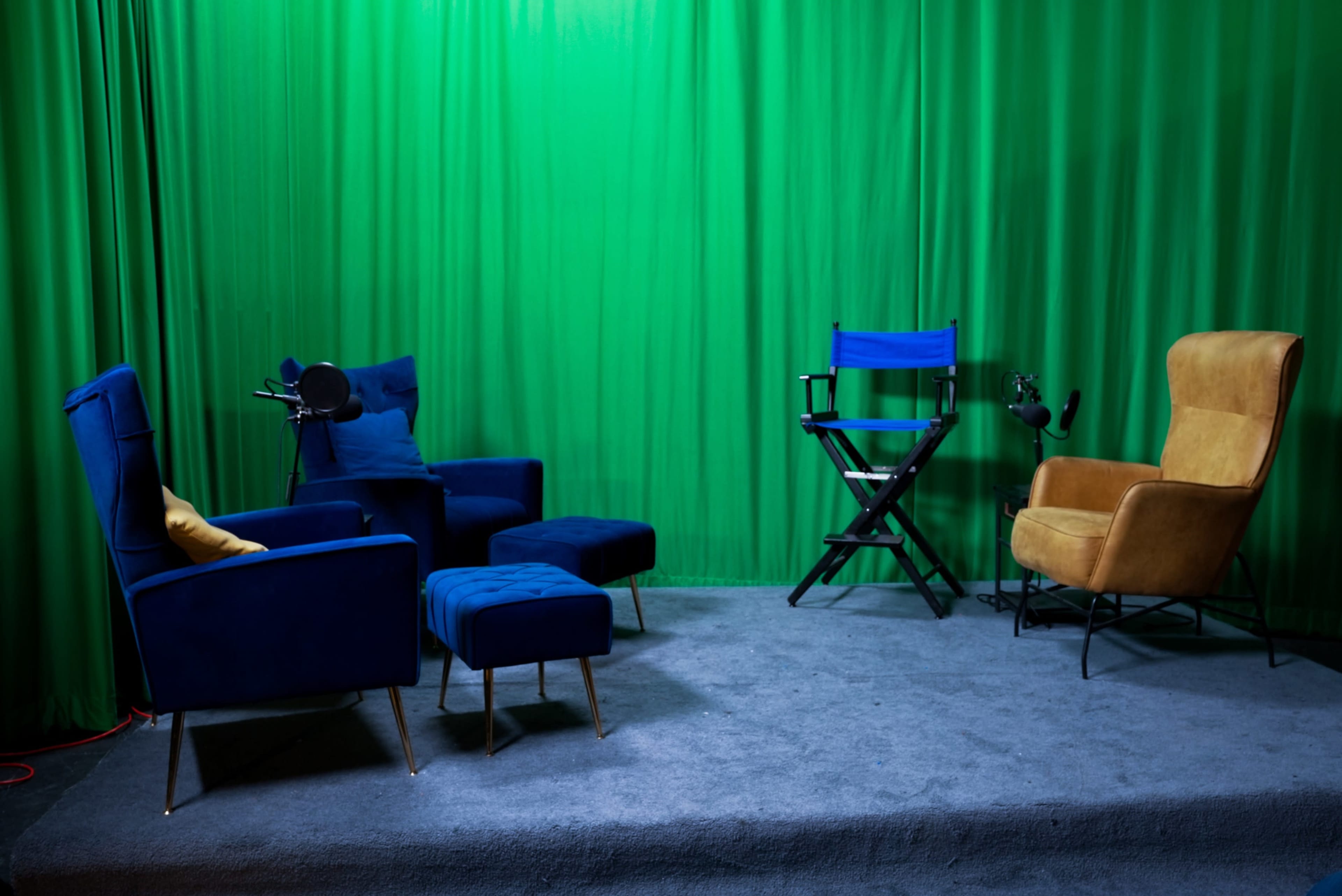 The image shows a small studio setup with blue and tan chairs, a director's chair, and a green backdrop.