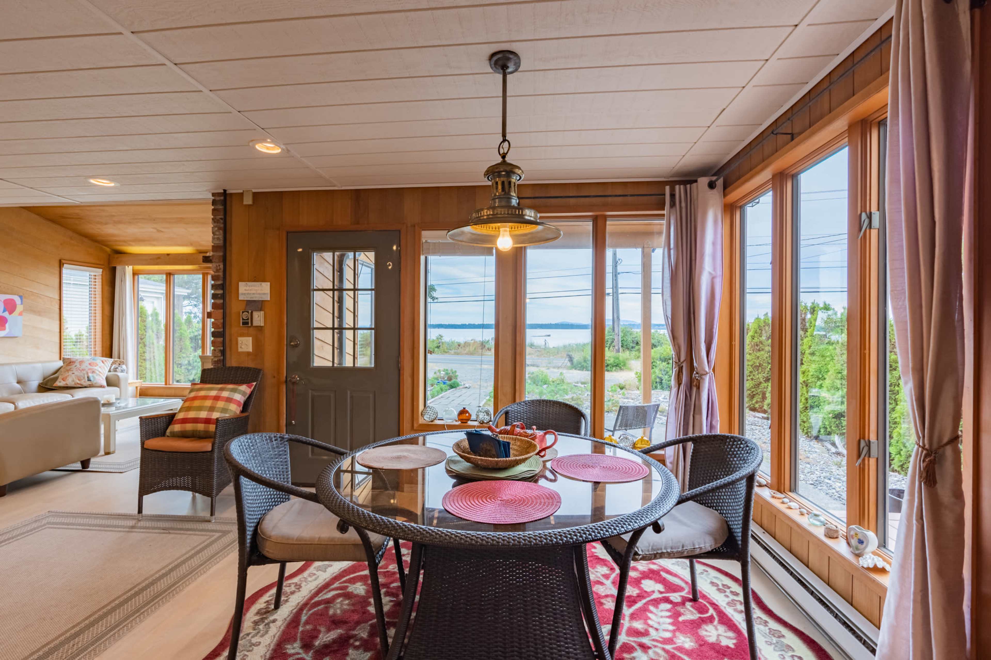 A glass-topped dining table with wicker chairs is set in a room with large windows providing a view of a body of water.