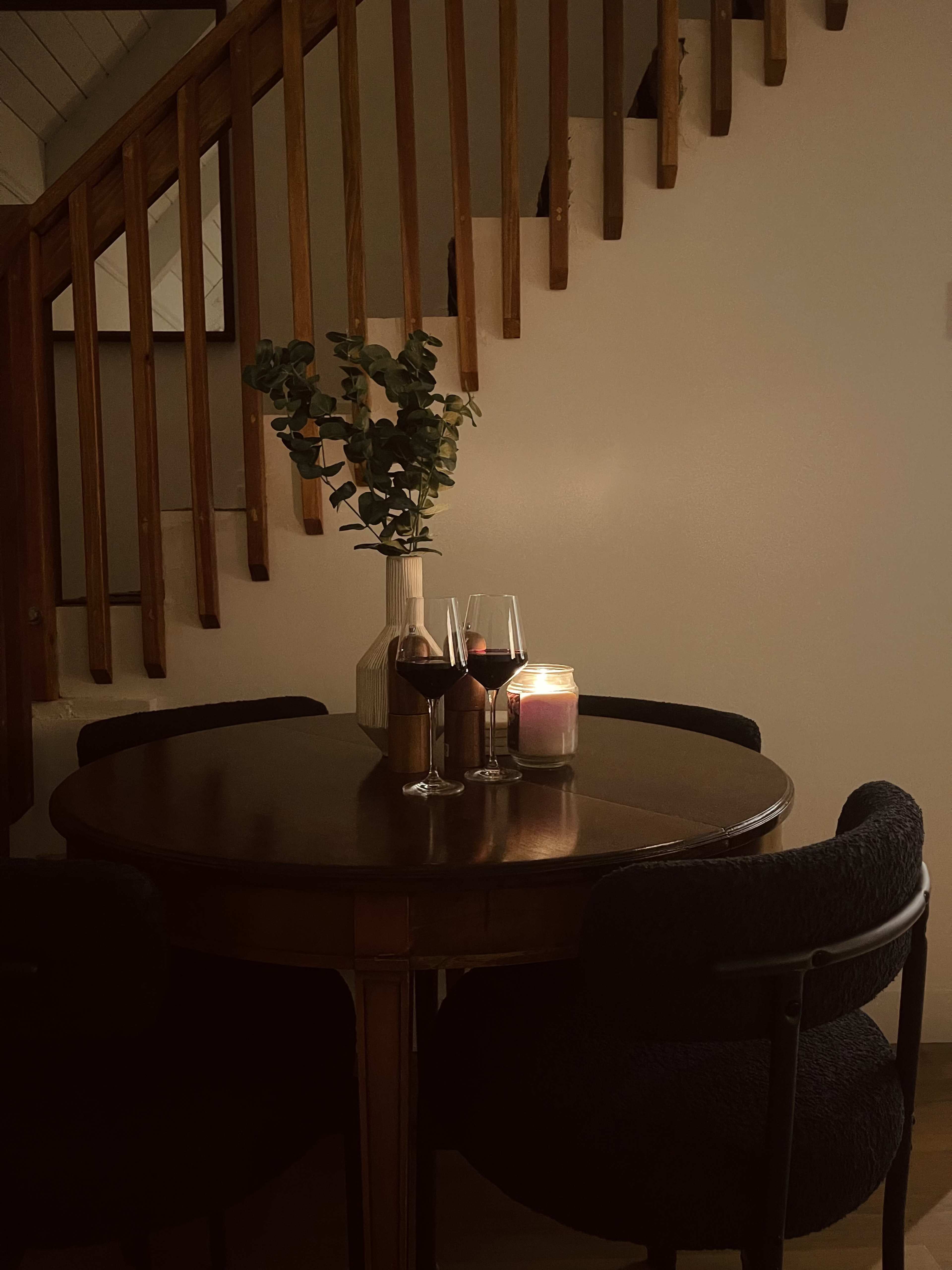 A wooden table is set with two glasses of red wine and a lit candle, surrounded by dark chairs and a staircase in the background.