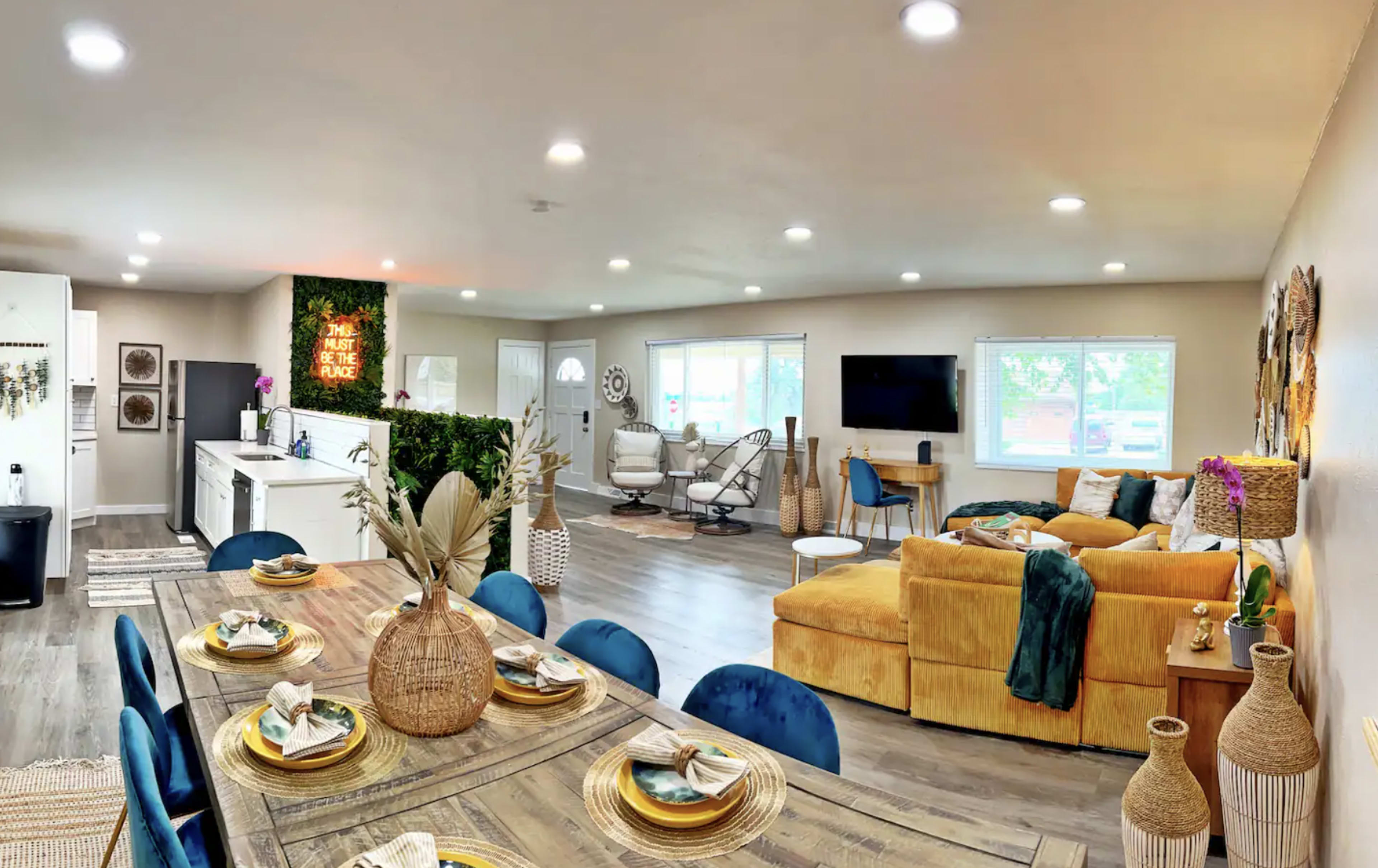 A modern, open-concept living space with a dining area featuring a wooden table and blue chairs, a well-lit kitchen, and a cozy seating area with a yellow couch.
