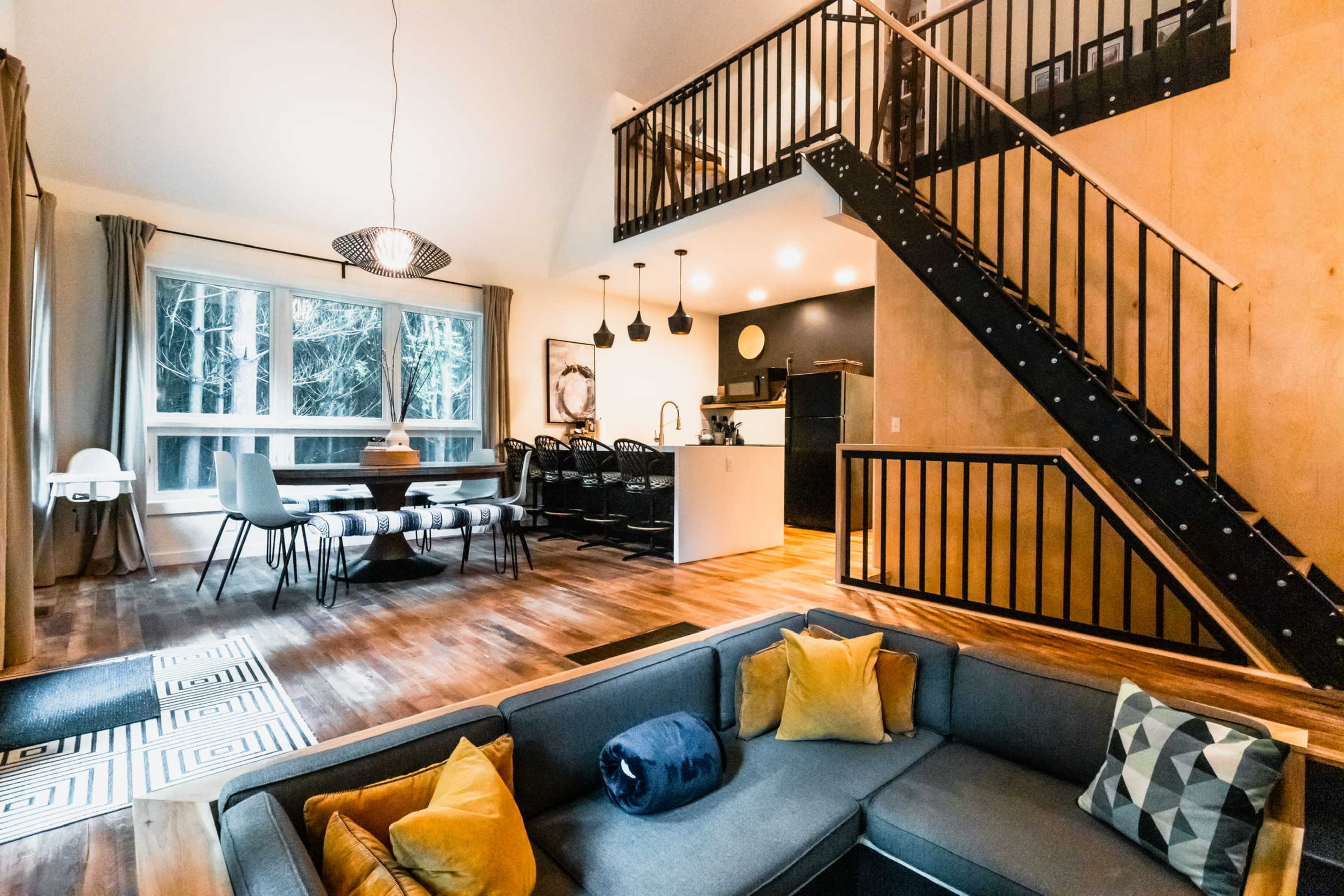 A modern living space with a sectional sofa in the foreground, a dining area, and an open kitchen, all featuring large windows that overlook a wooded landscape.