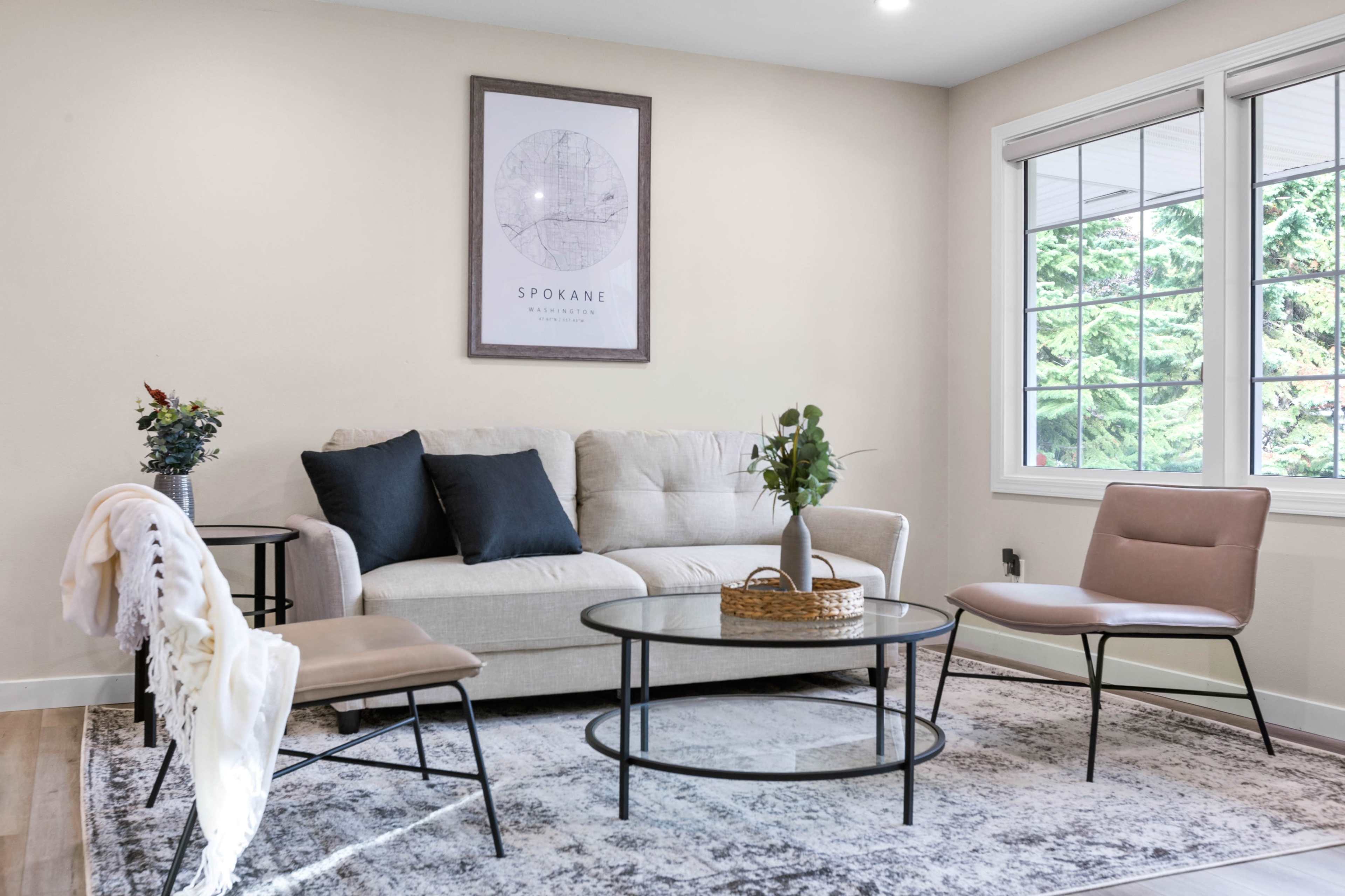 A light-colored living room features a beige sofa, two chairs, a round coffee table, and a framed map on the wall.