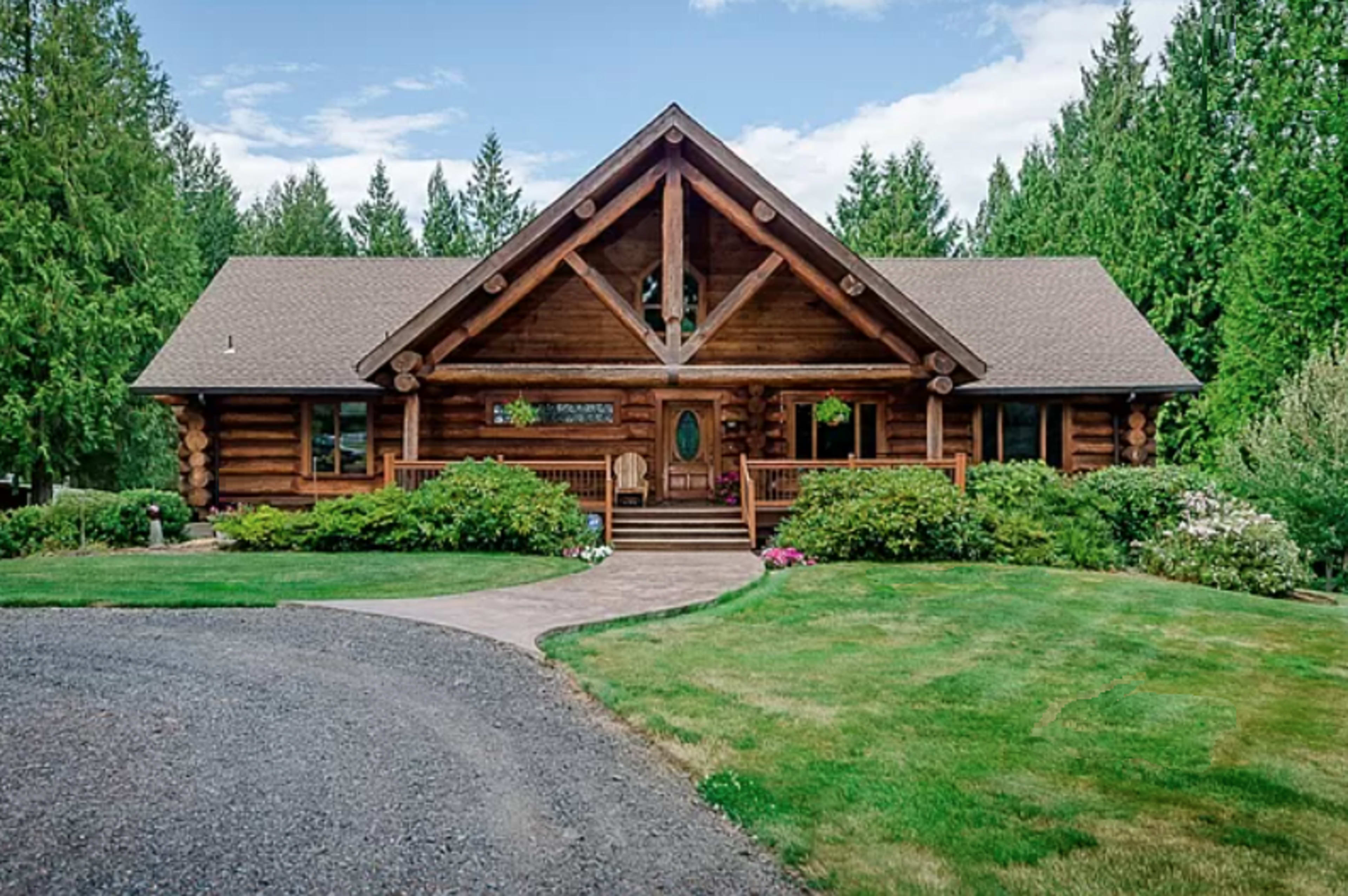 A log cabin-style house sits amid green trees and landscaped gardens, featuring a front porch and a stone walkway.