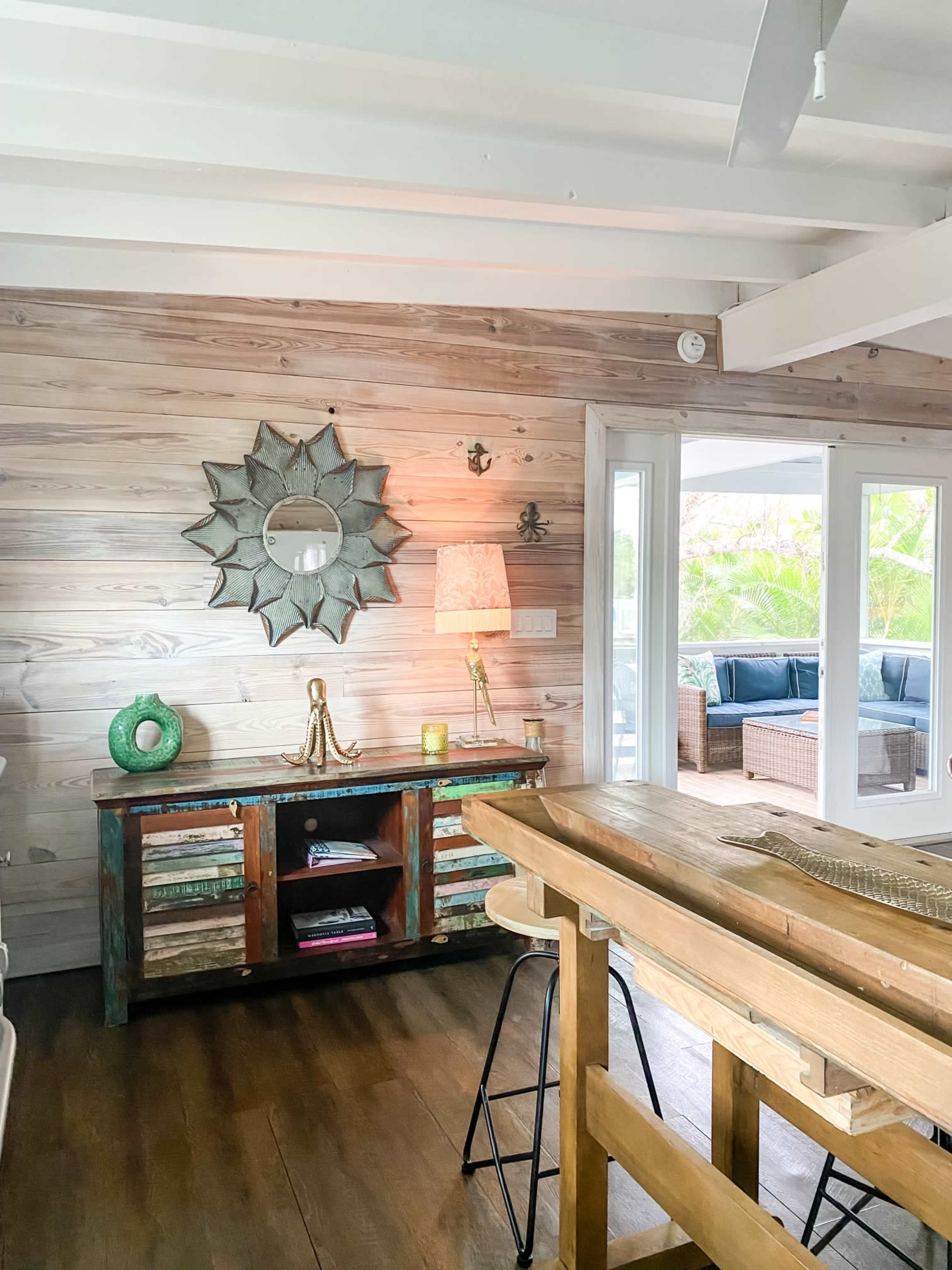 The image shows a wooden interior with a decorative mirror on the wall, a lamp on a rustic side table, and a view of a seating area through an open door.