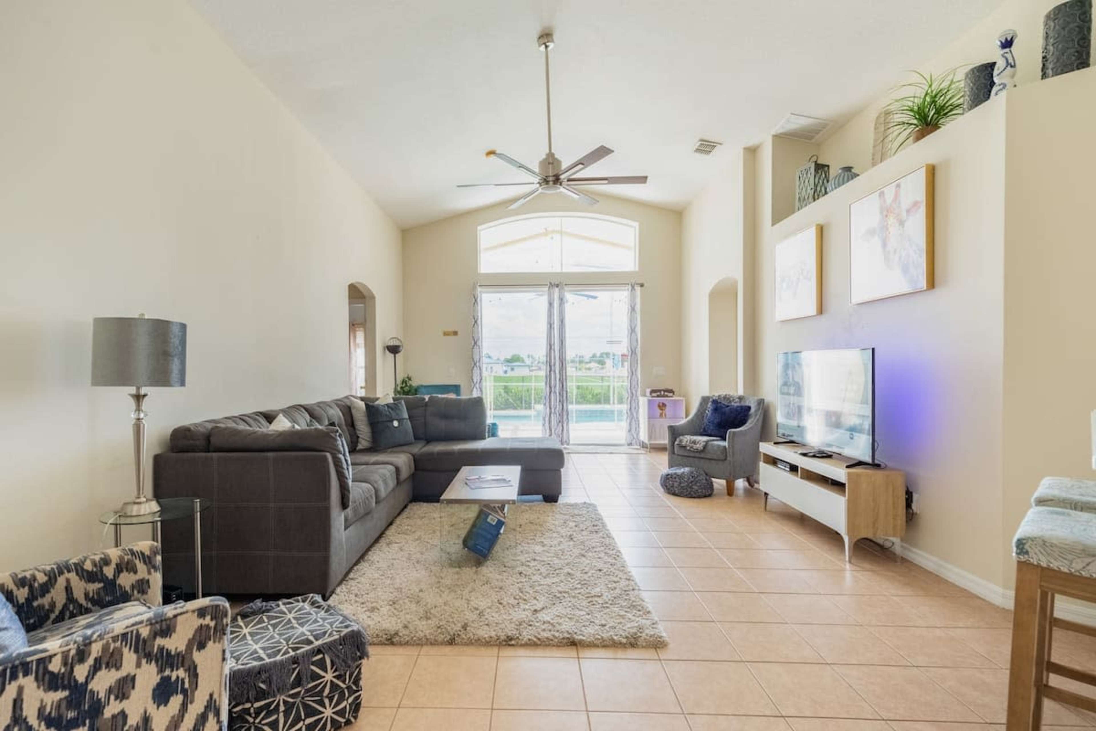 The image shows a living room with a large sectional couch, a coffee table, and a television, featuring tile flooring and large windows that allow natural light.