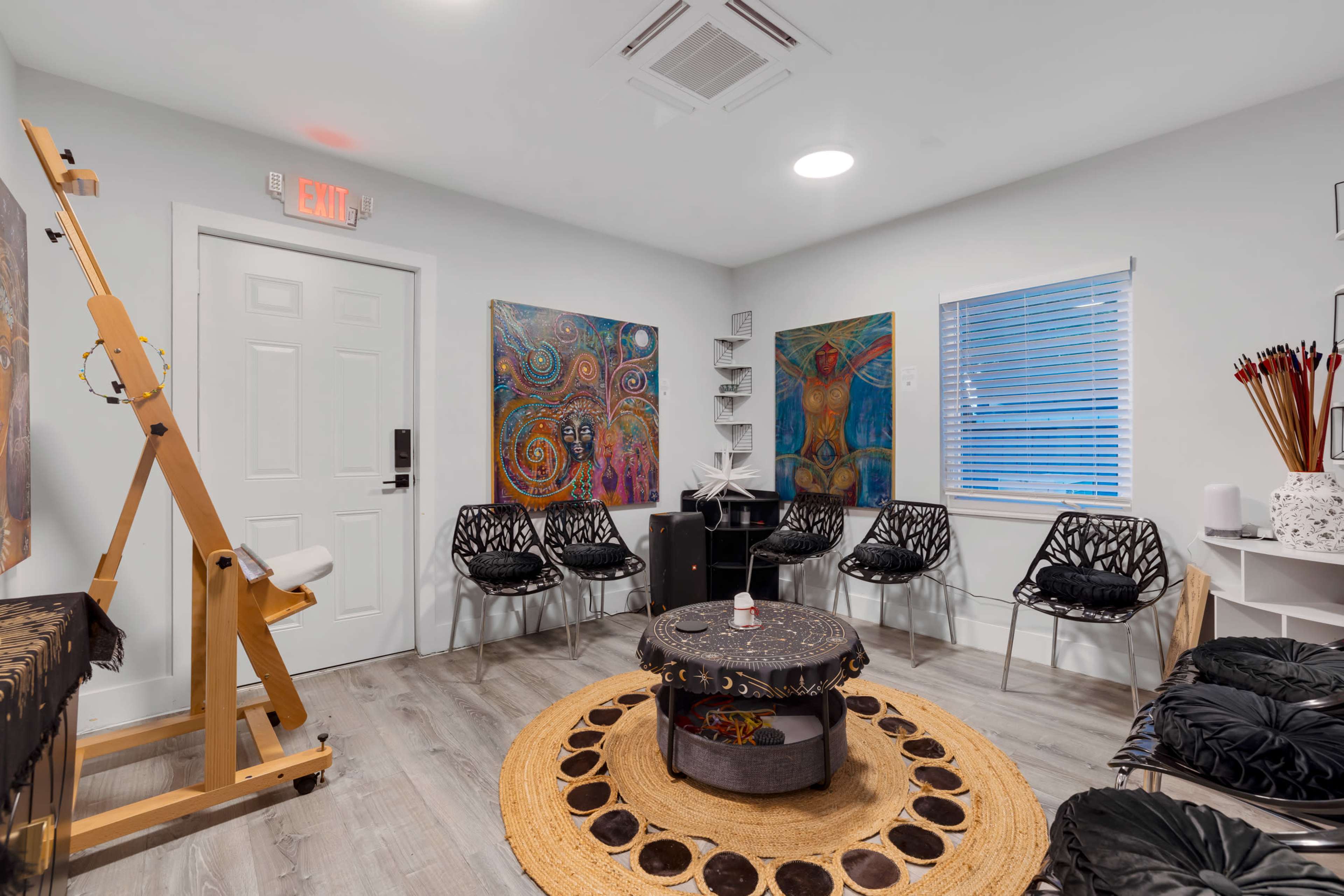 The image shows a small, well-lit room with six black chairs arranged around a circular table, decorated with colorful artworks on the walls and an easel in one corner.