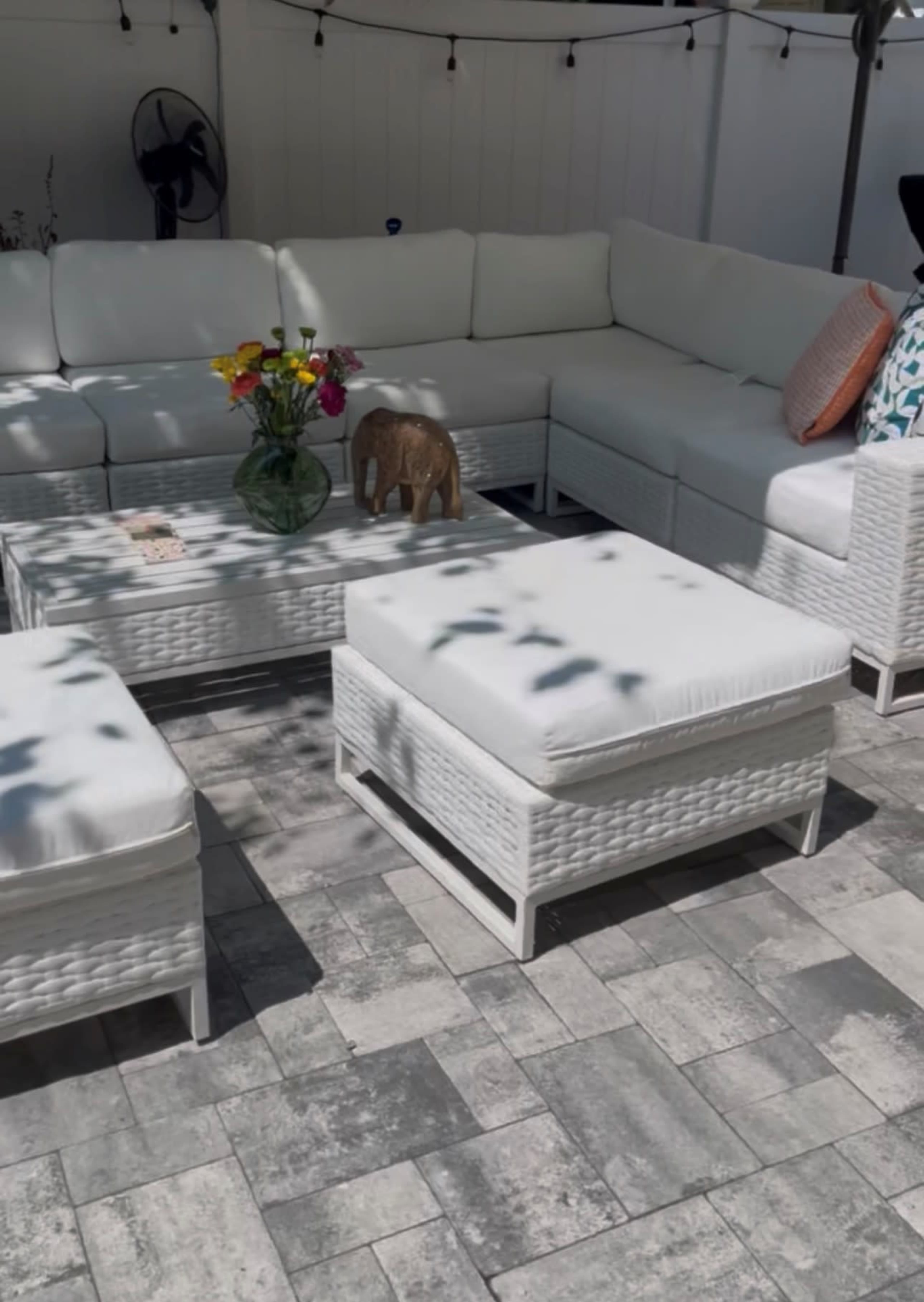 The image shows a cozy outdoor seating area with white wicker furniture, a coffee table, and a vase of flowers on top, all set on a stone patio.