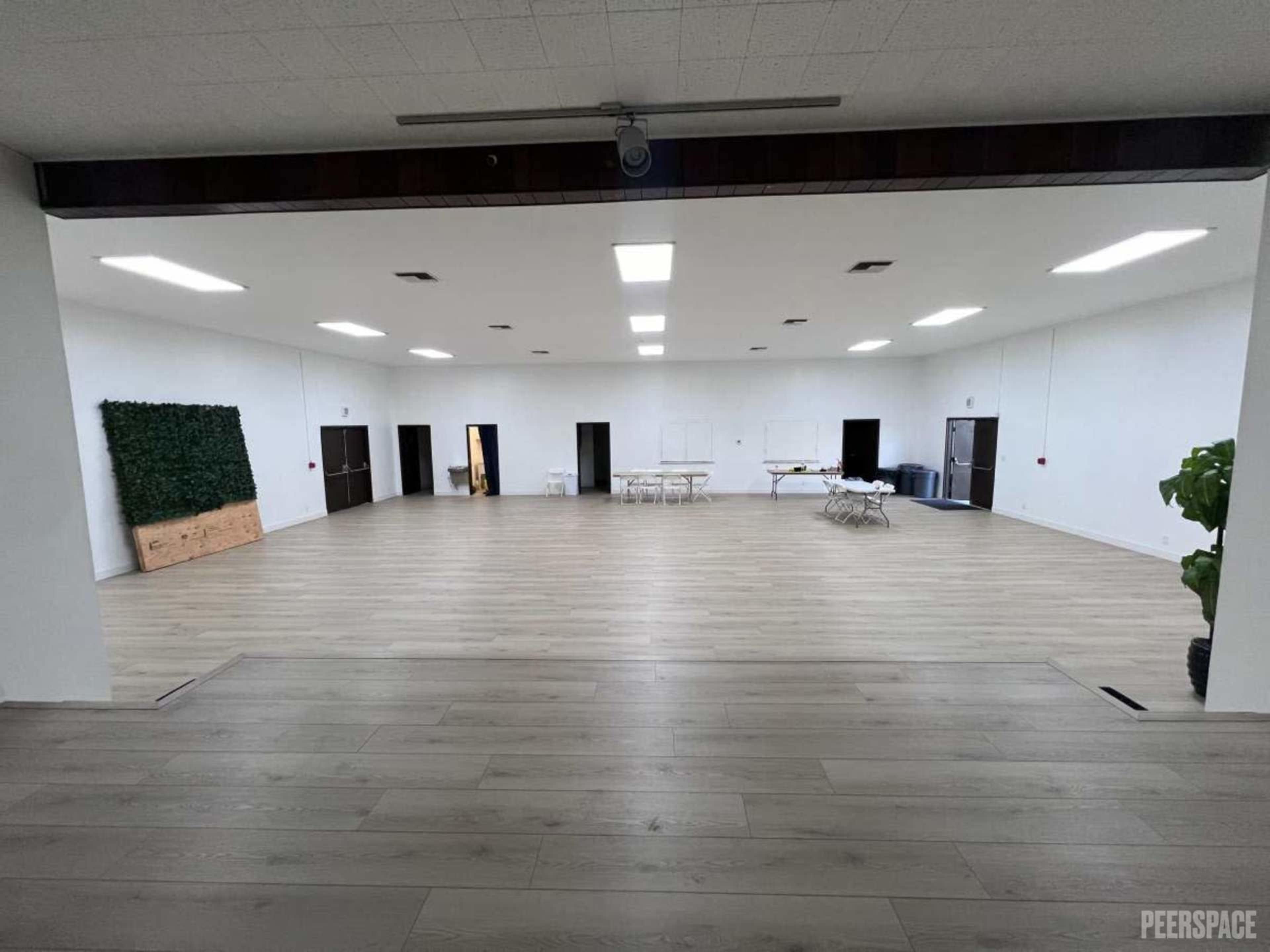 The image shows a spacious, empty interior room with wooden flooring, bright overhead lights, and simple furnishings along the walls.