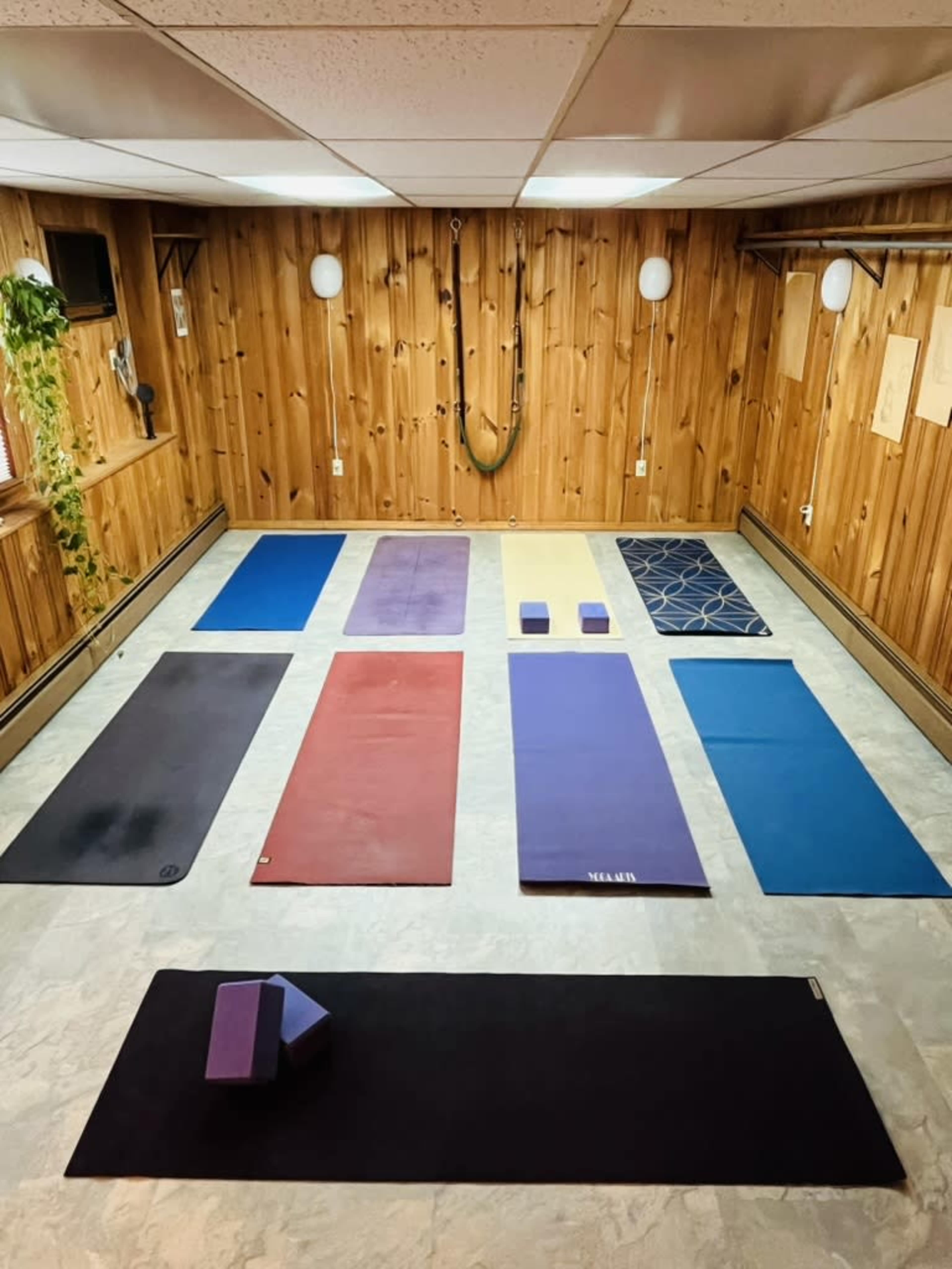 The image shows a yoga studio with several exercise mats of various colors arranged on a light-colored floor, surrounded by wooden paneling and minimal decor.