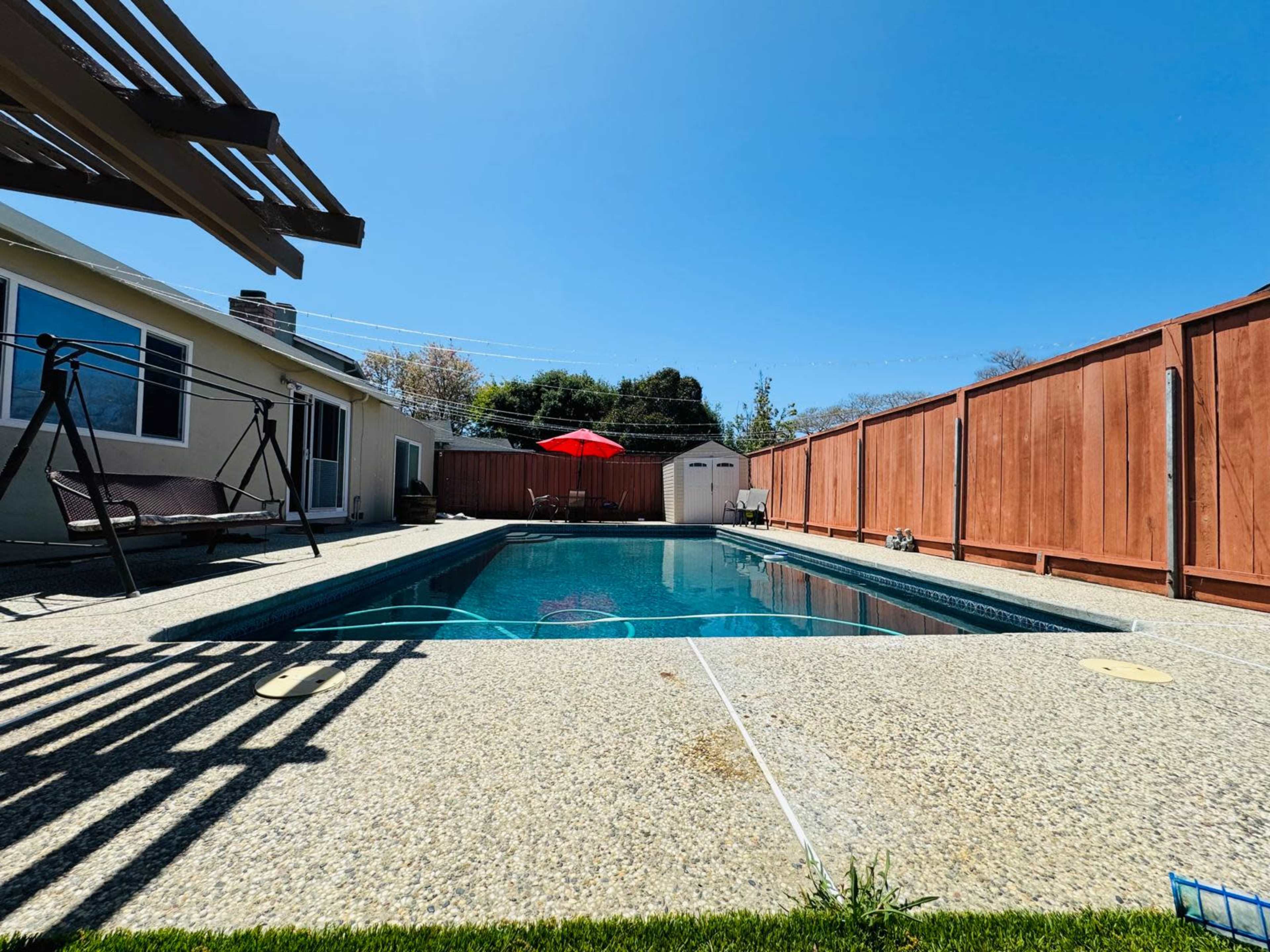 A swimming pool surrounded by a concrete patio, with a red umbrella providing shade and a wooden fence enclosing the area.