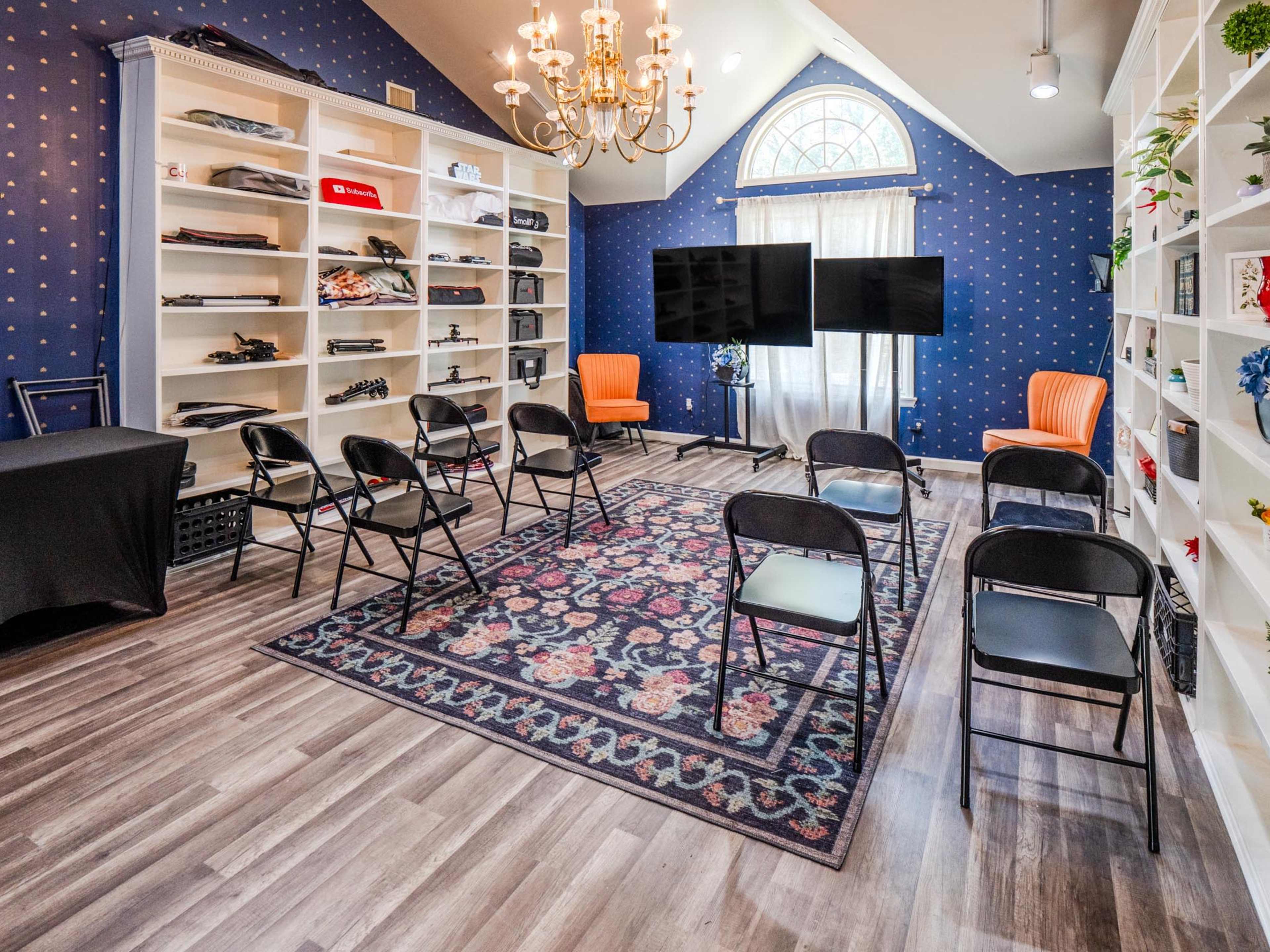 A well-lit room with blue wallpaper, a chandelier, shelves filled with items, two televisions on a stand, and chairs arranged in a semi-circle on a floral rug.