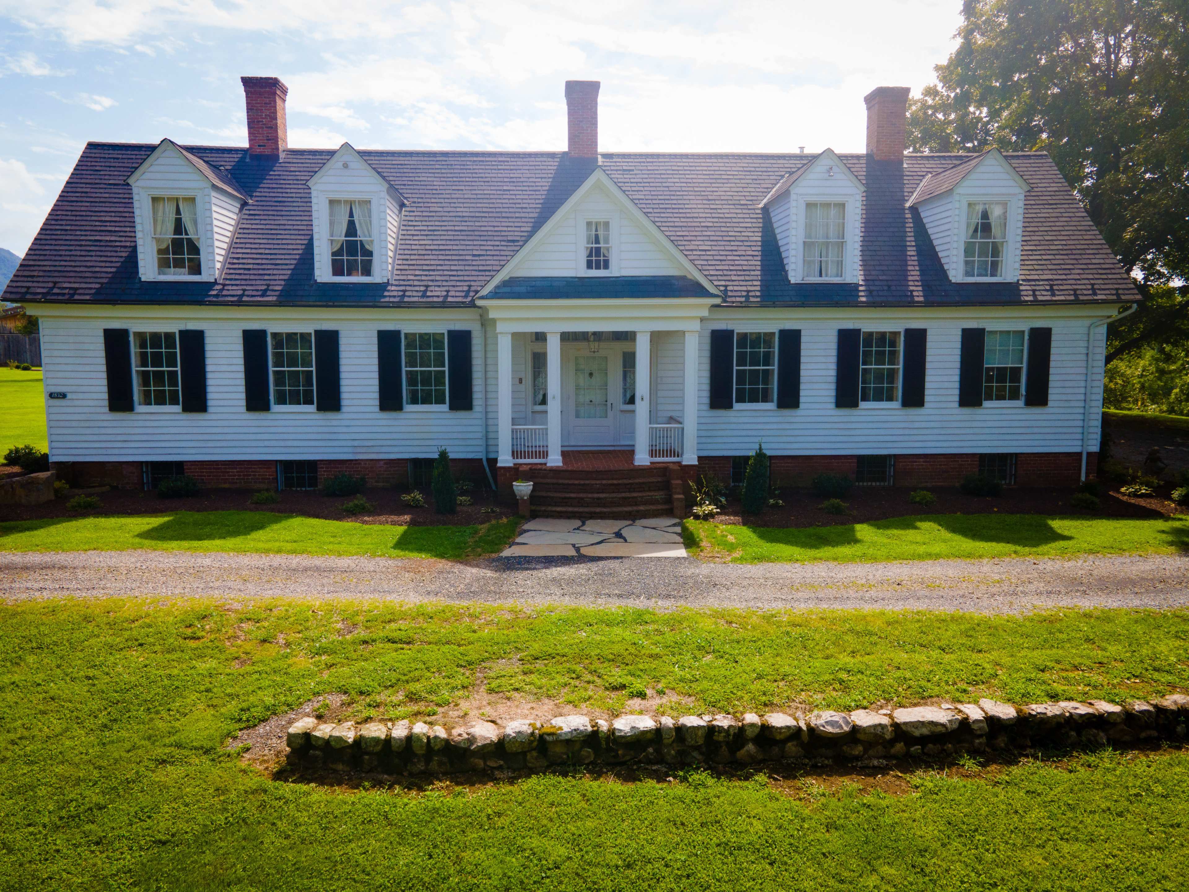 A two-story white house with a gabled roof, black shutters, and a front porch is situated on a green lawn, bordered by a stone wall.