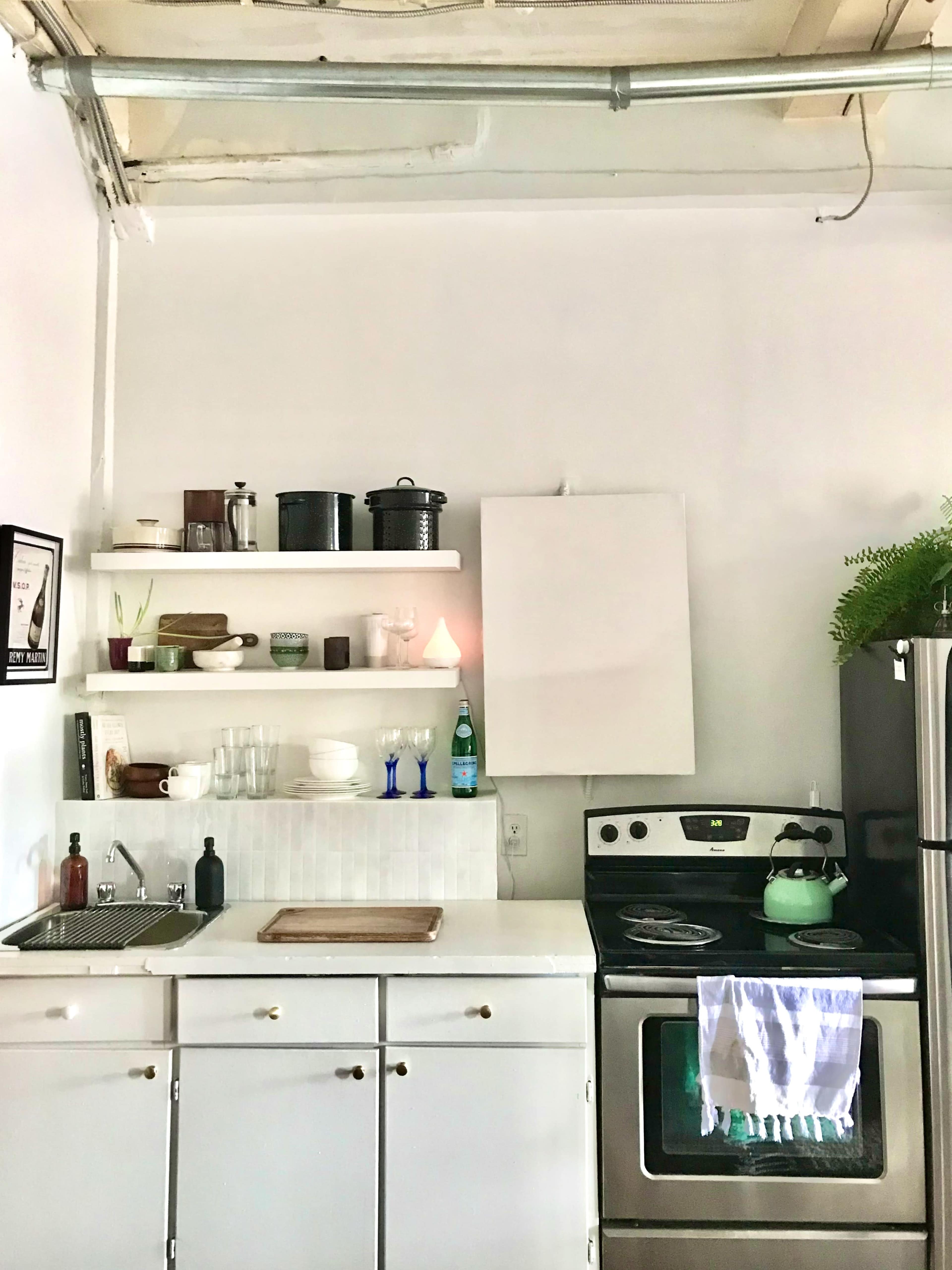 A compact kitchen features white cabinets, a stainless steel stove, wall shelves with various kitchen items, and a small sink.