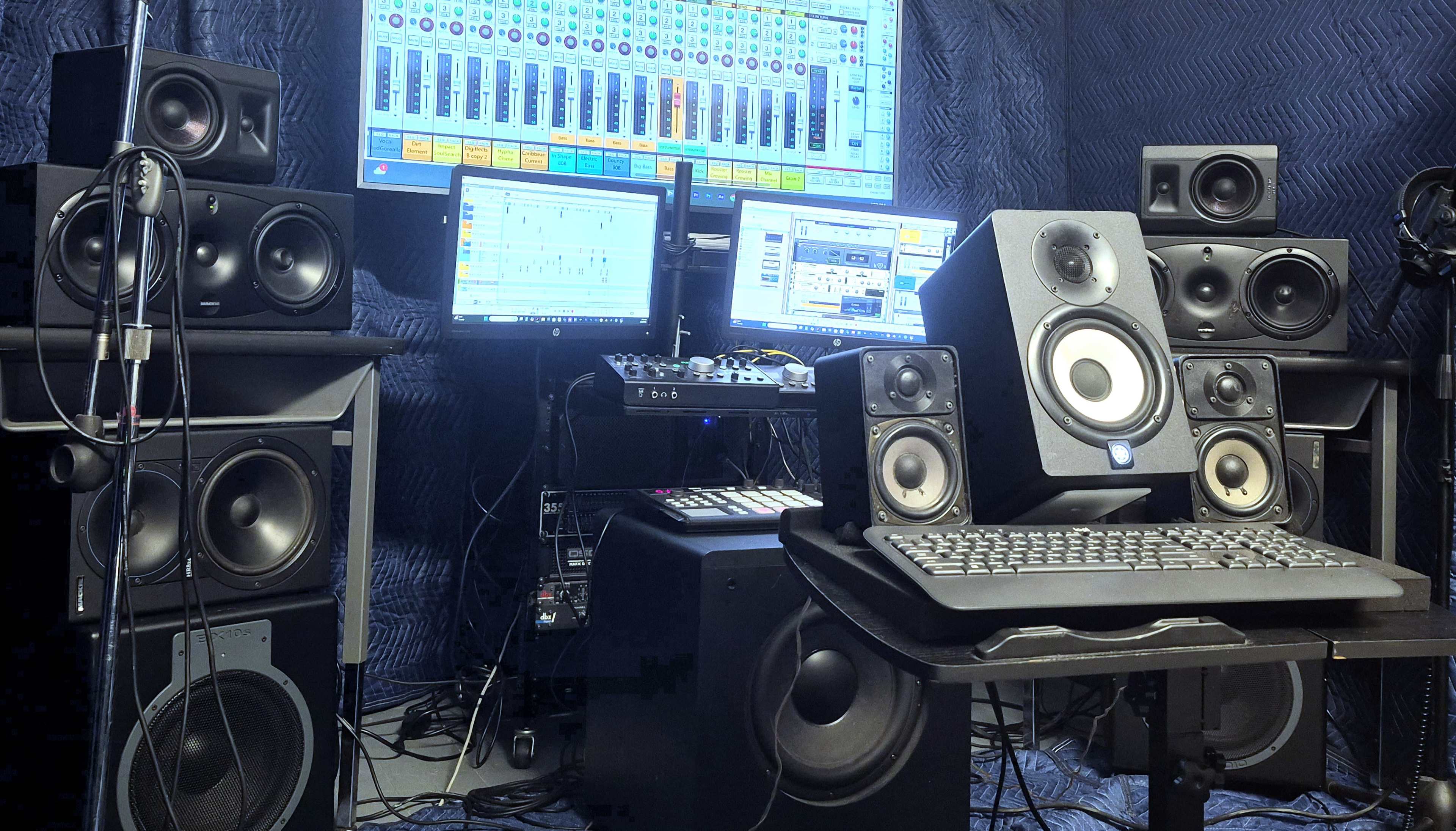 The image shows a music production setup featuring multiple speakers, a computer with two monitors, a MIDI controller, and various audio equipment arranged on a desk.