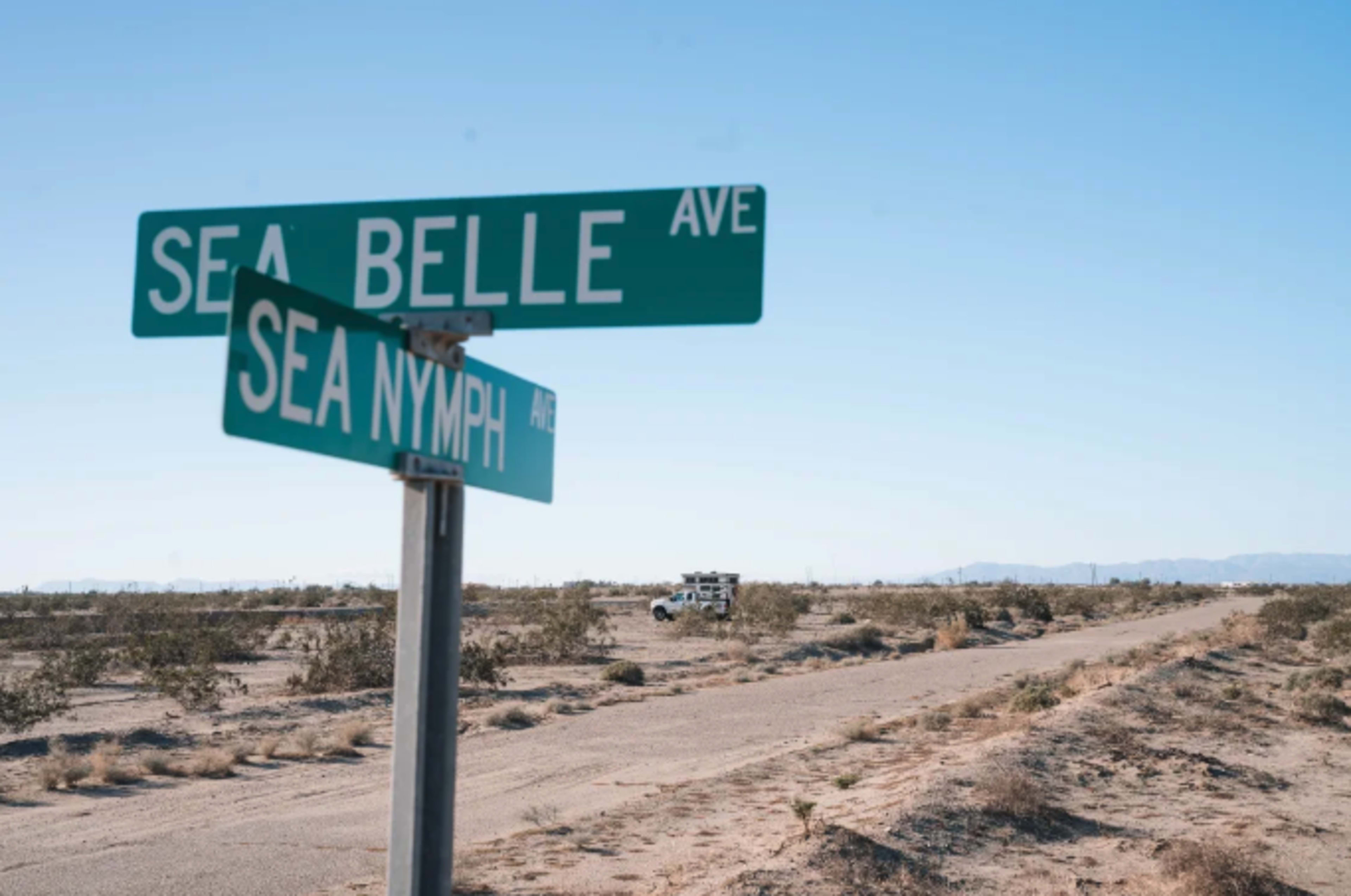 A street sign at the intersection of Sea Belle Avenue and Sea Nymph Avenue stands in a desert landscape with a vehicle visible in the distance.