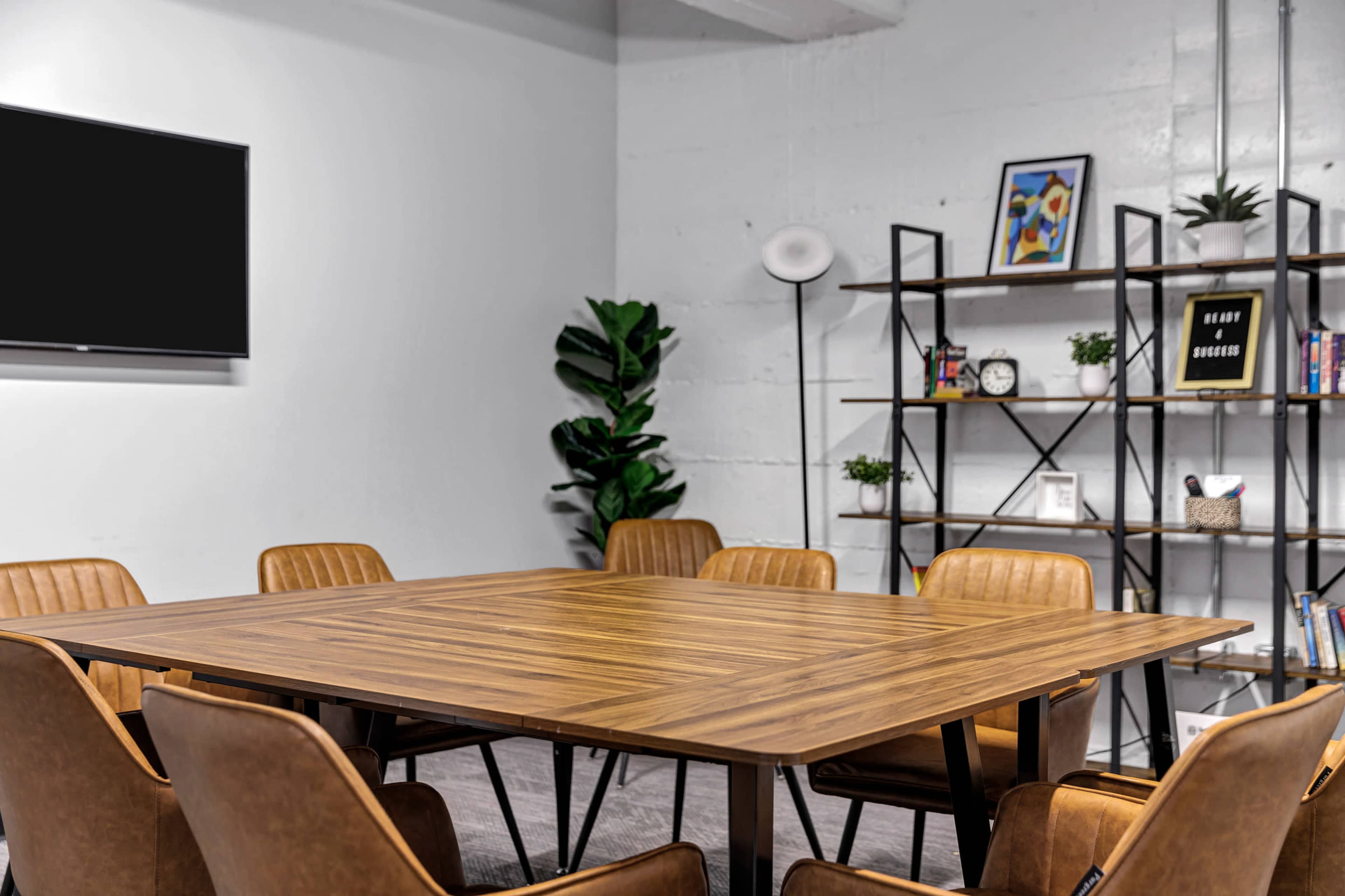 A meeting room features a wooden table surrounded by brown leather chairs, with a TV on the wall and a shelving unit displaying decorative items.