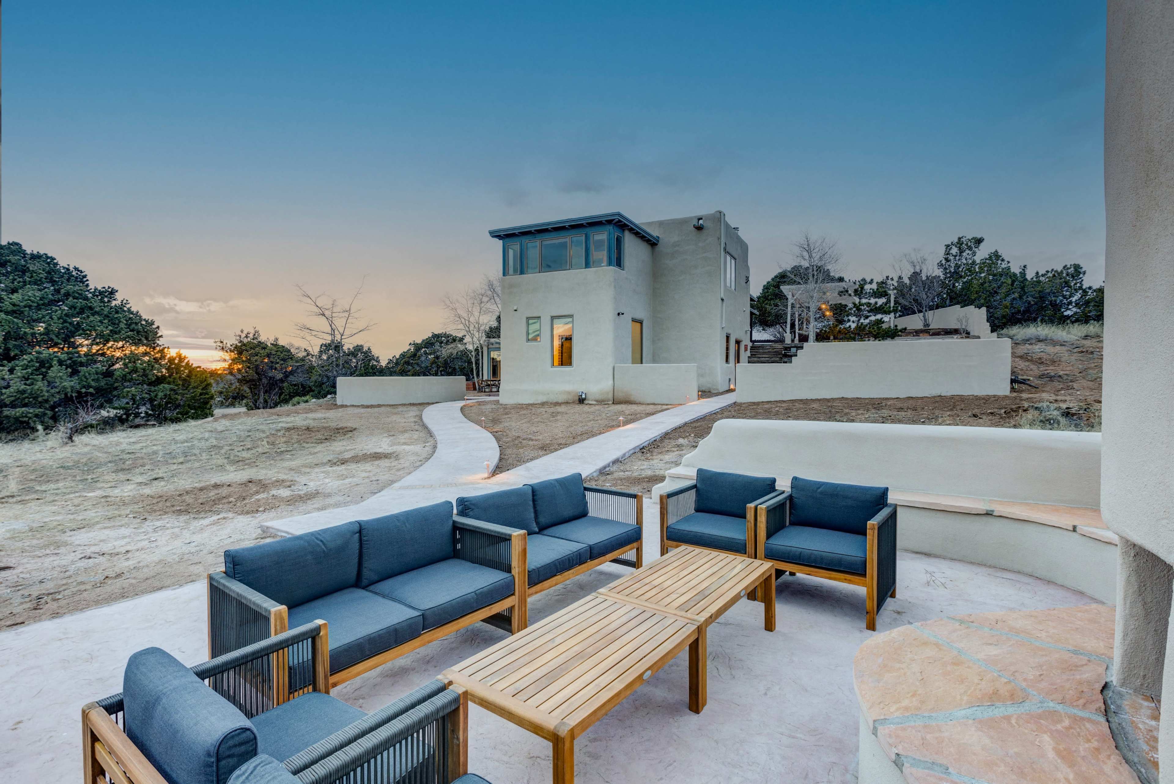 A modern house sits on elevated ground with a pathway leading to it, surrounded by sparse vegetation and outdoor seating in the foreground.