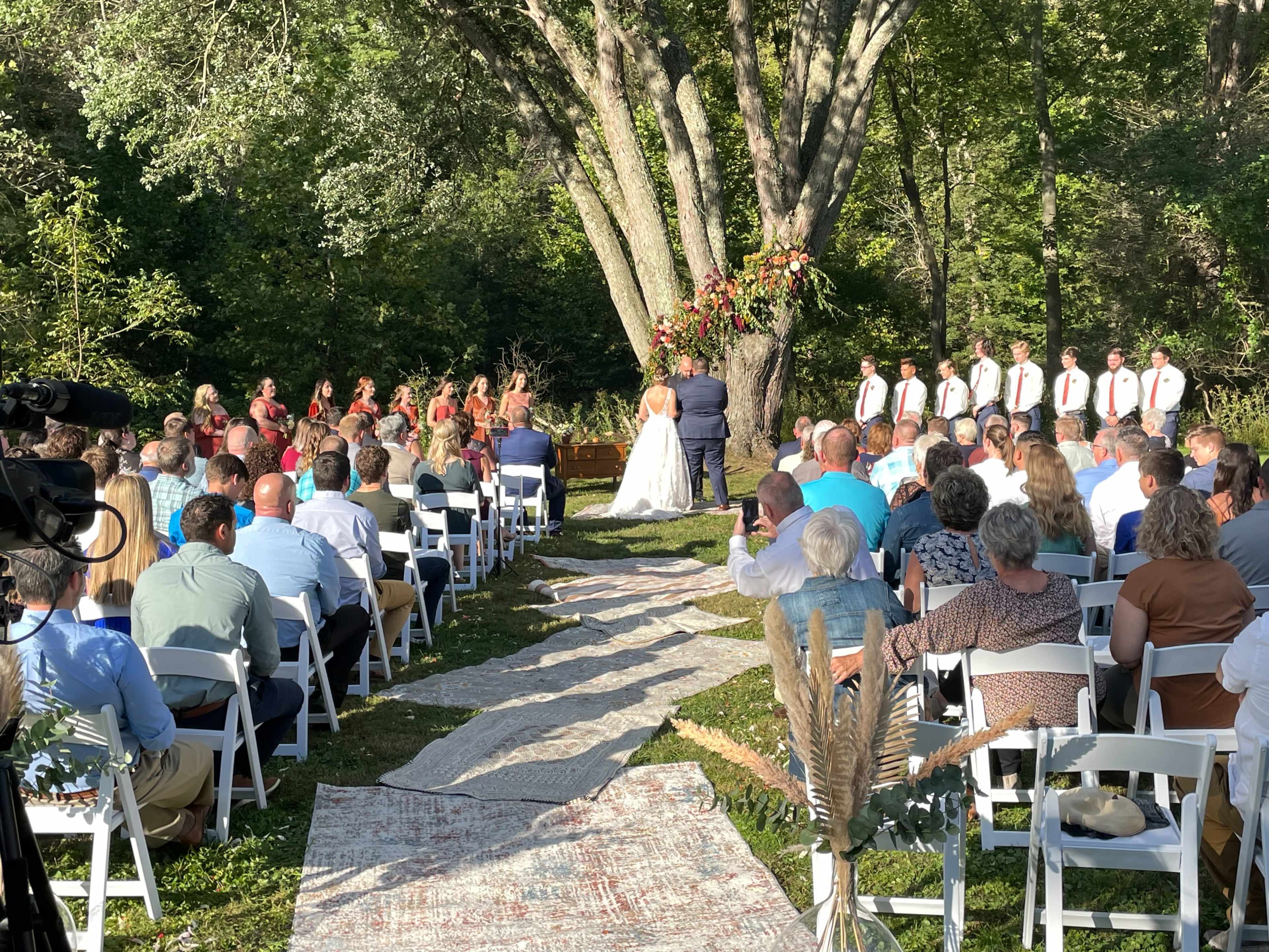 A wedding ceremony takes place outdoors under a tree, with guests seated on white chairs and a decorated aisle leading to the couple at the altar.