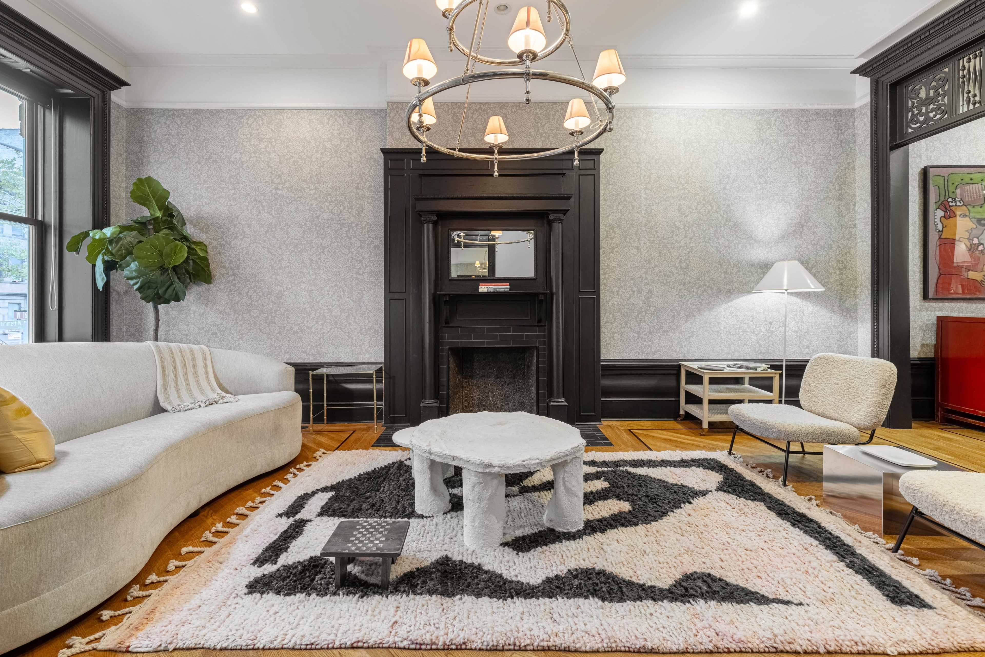 A living room featuring a plush gray sofa, a large patterned area rug, a marble coffee table, and a striking chandelier above an elegant fireplace.