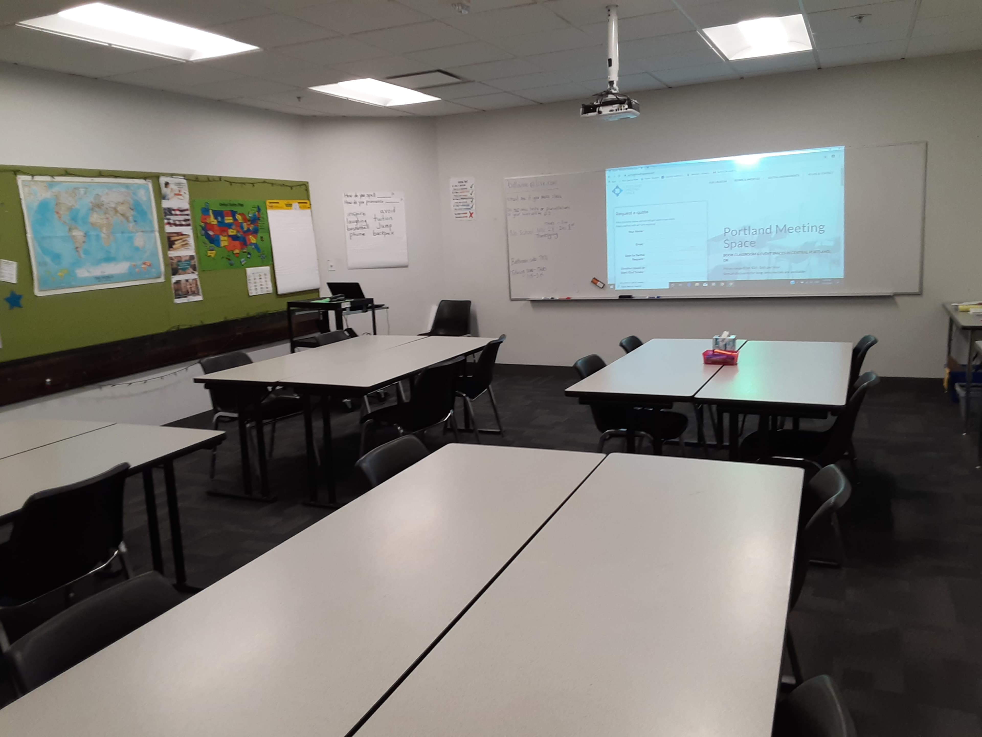 A classroom is arranged with several tables and chairs, featuring a projector and whiteboard on one wall along with a map.
