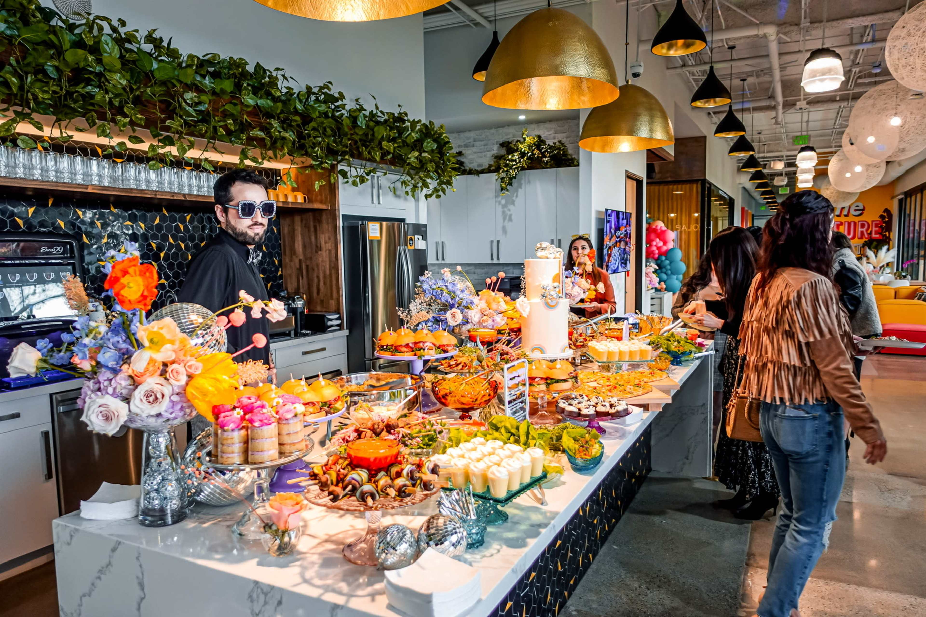 A buffet table filled with an array of colorful dishes and desserts is set up in a modern, well-decorated space, with guests milling around and a person wearing sunglasses standing behind the table.