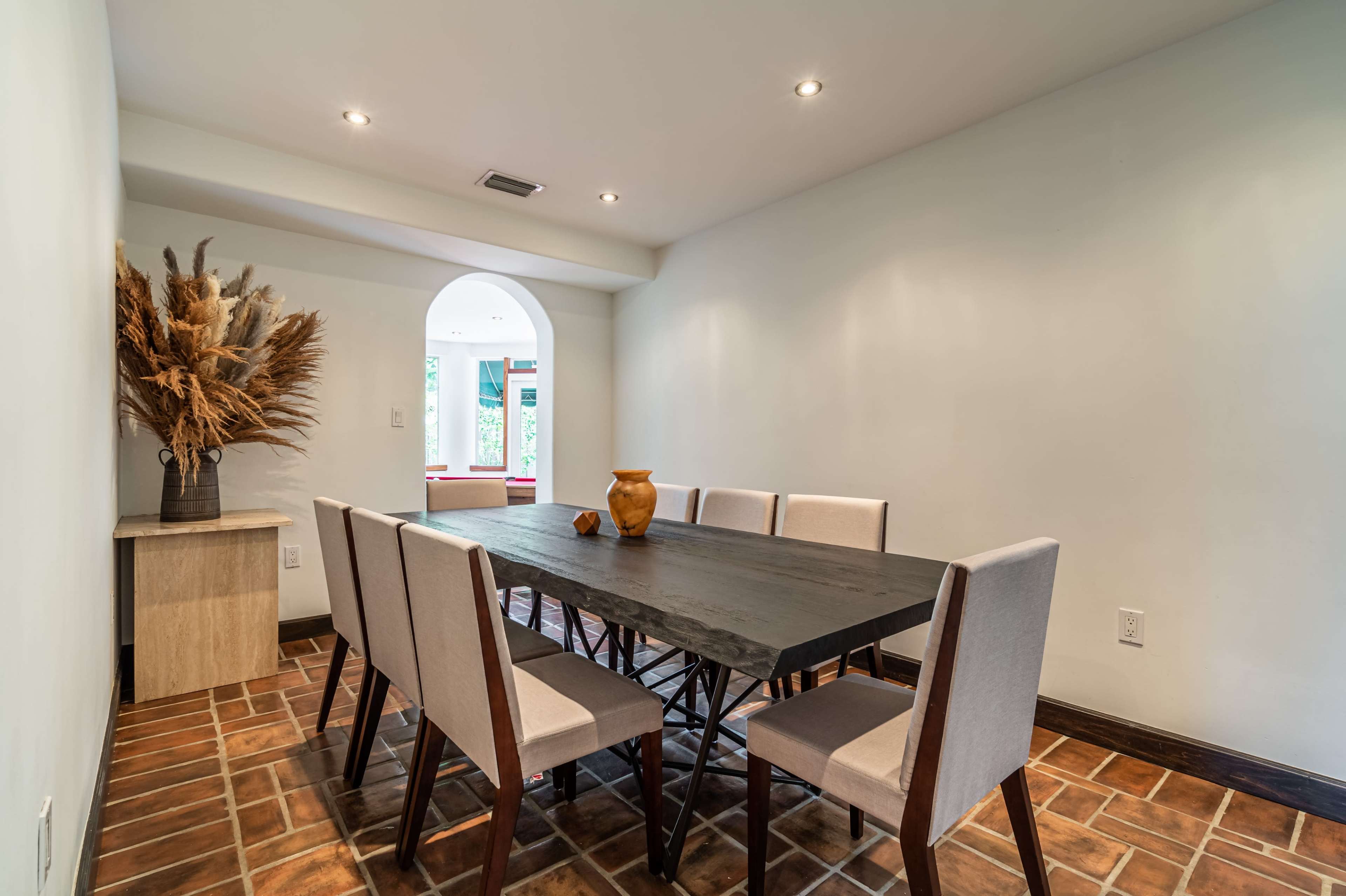 The image shows a dining room featuring a large black table surrounded by eight upholstered chairs and a decorative arrangement of dried plants in a corner.