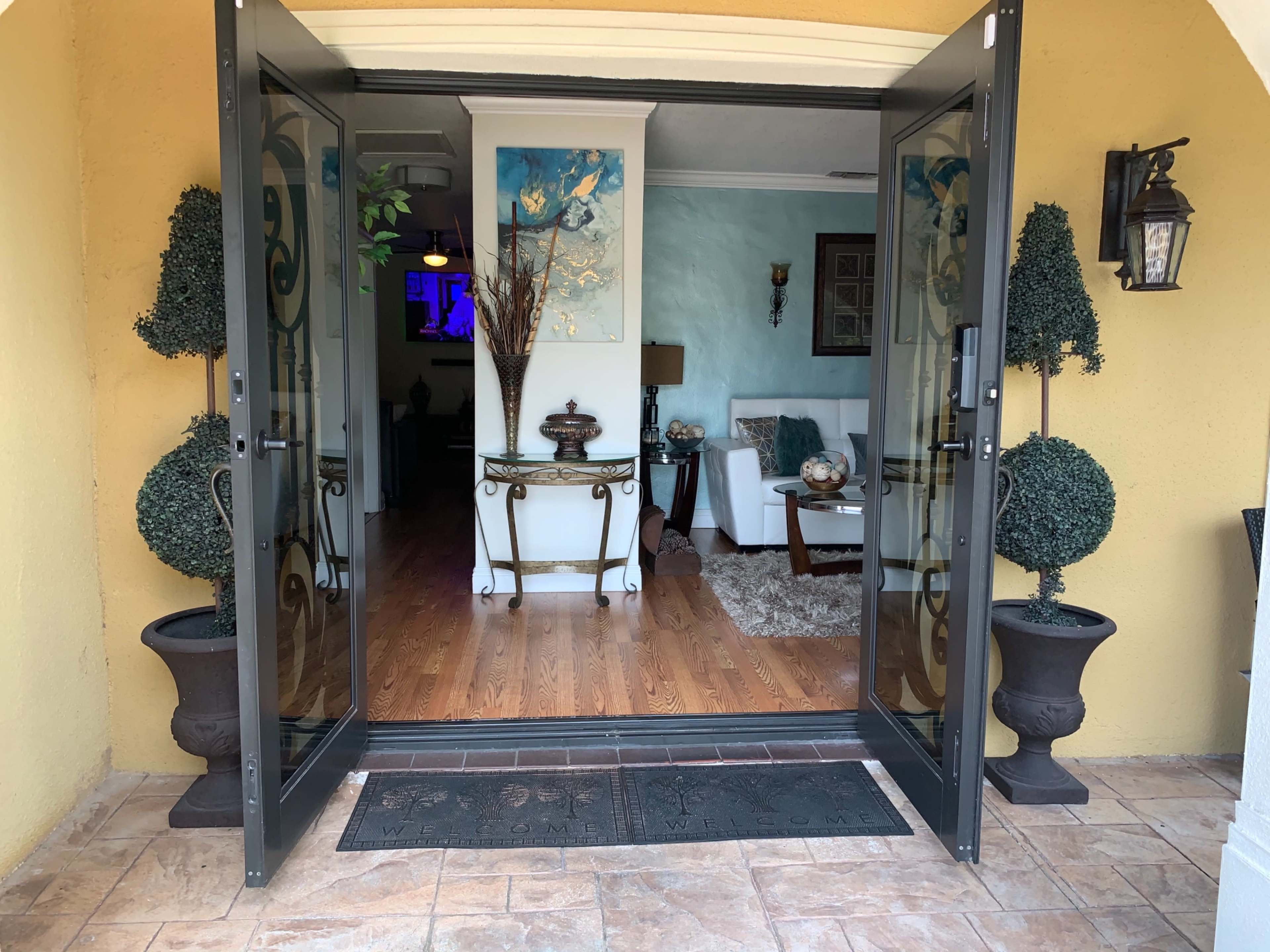 The image shows a pair of open glass doors leading into a well-decorated living room with wooden flooring and light-colored walls.
