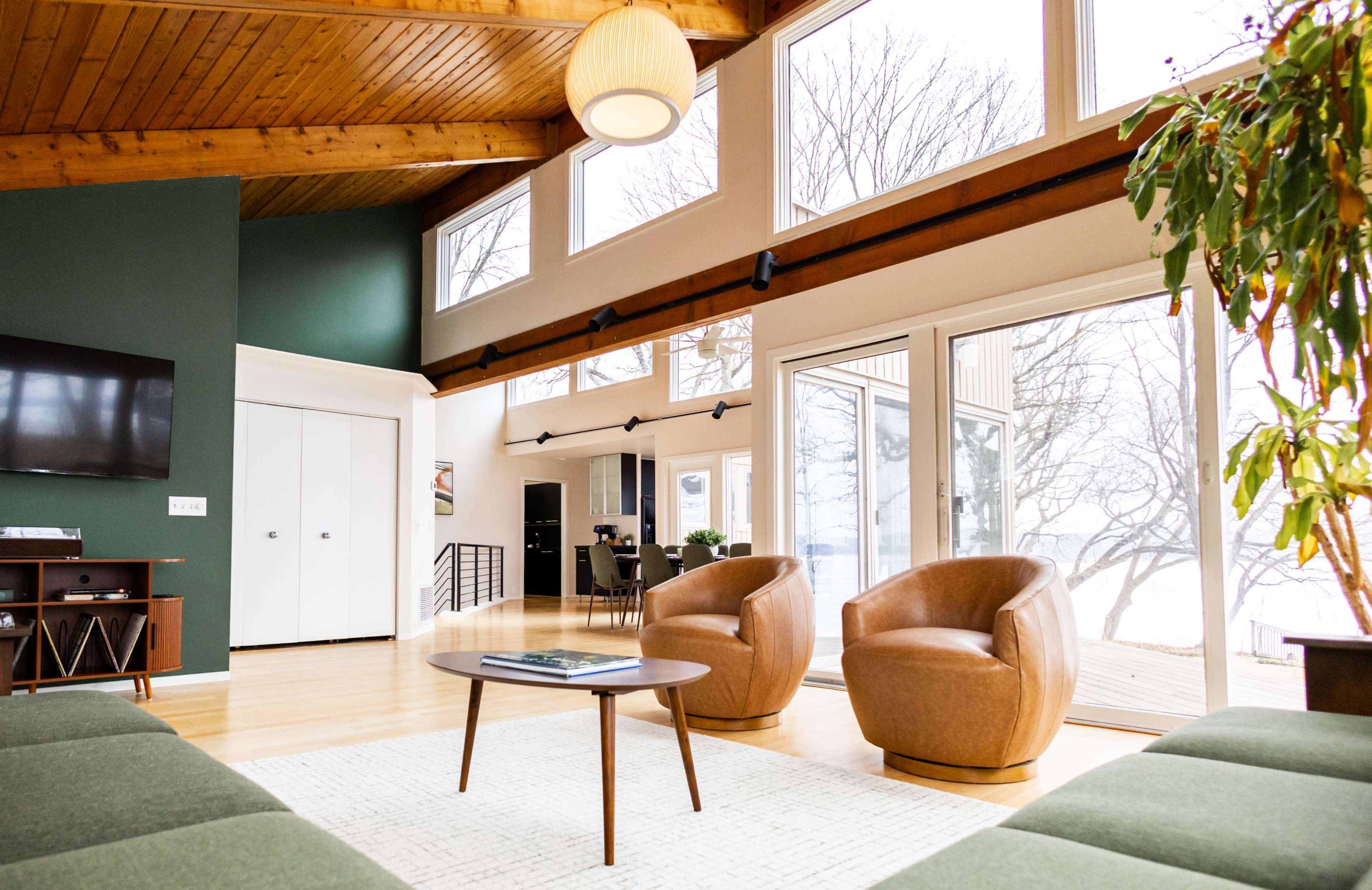 A spacious living area features large windows, a wooden ceiling, two brown armchairs, and a coffee table, providing a view of the outside landscape.