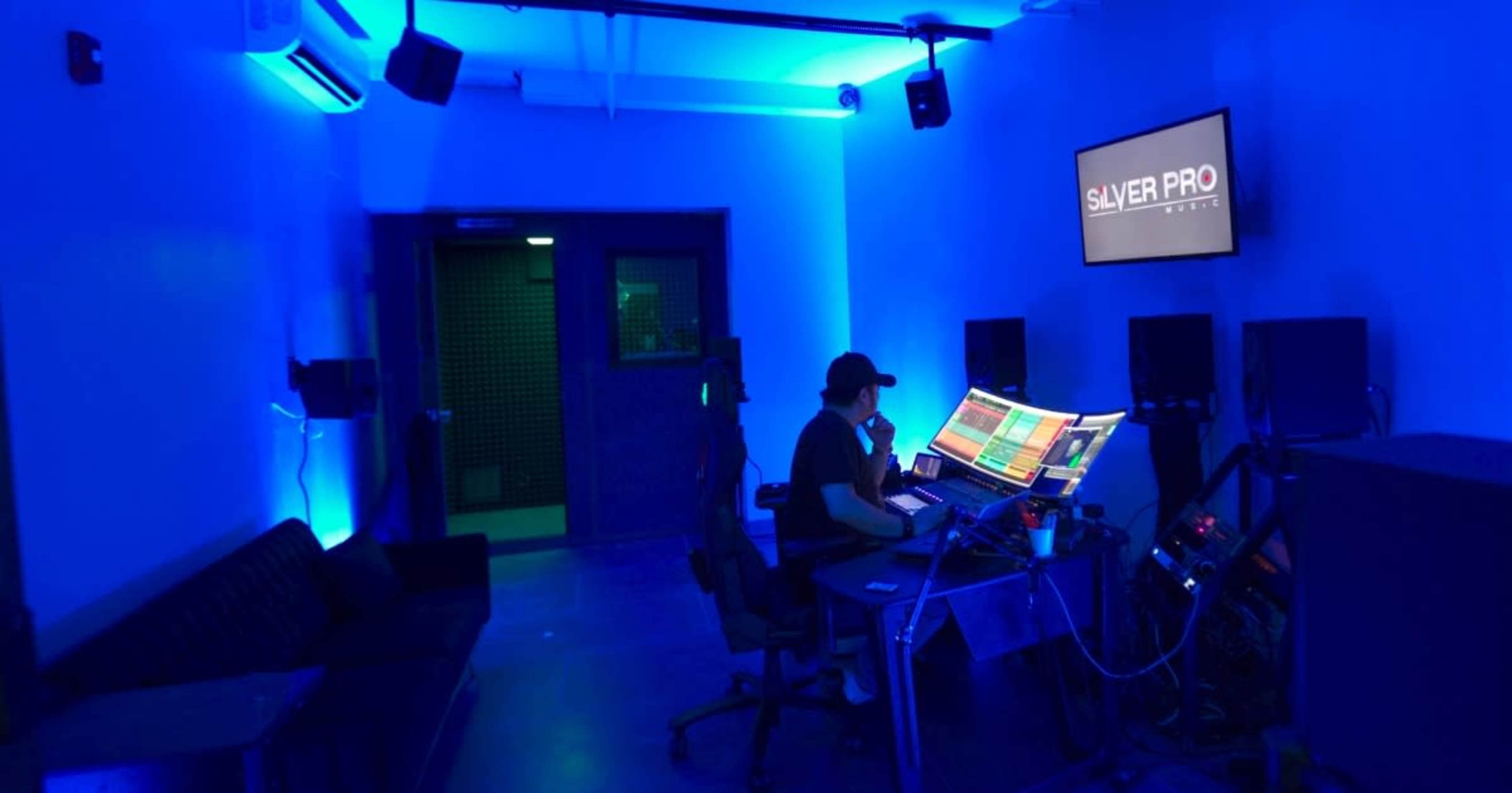 A person is sitting at a computer workstation in a dimly lit recording studio, with blue ambient lighting and sound equipment visible around them.