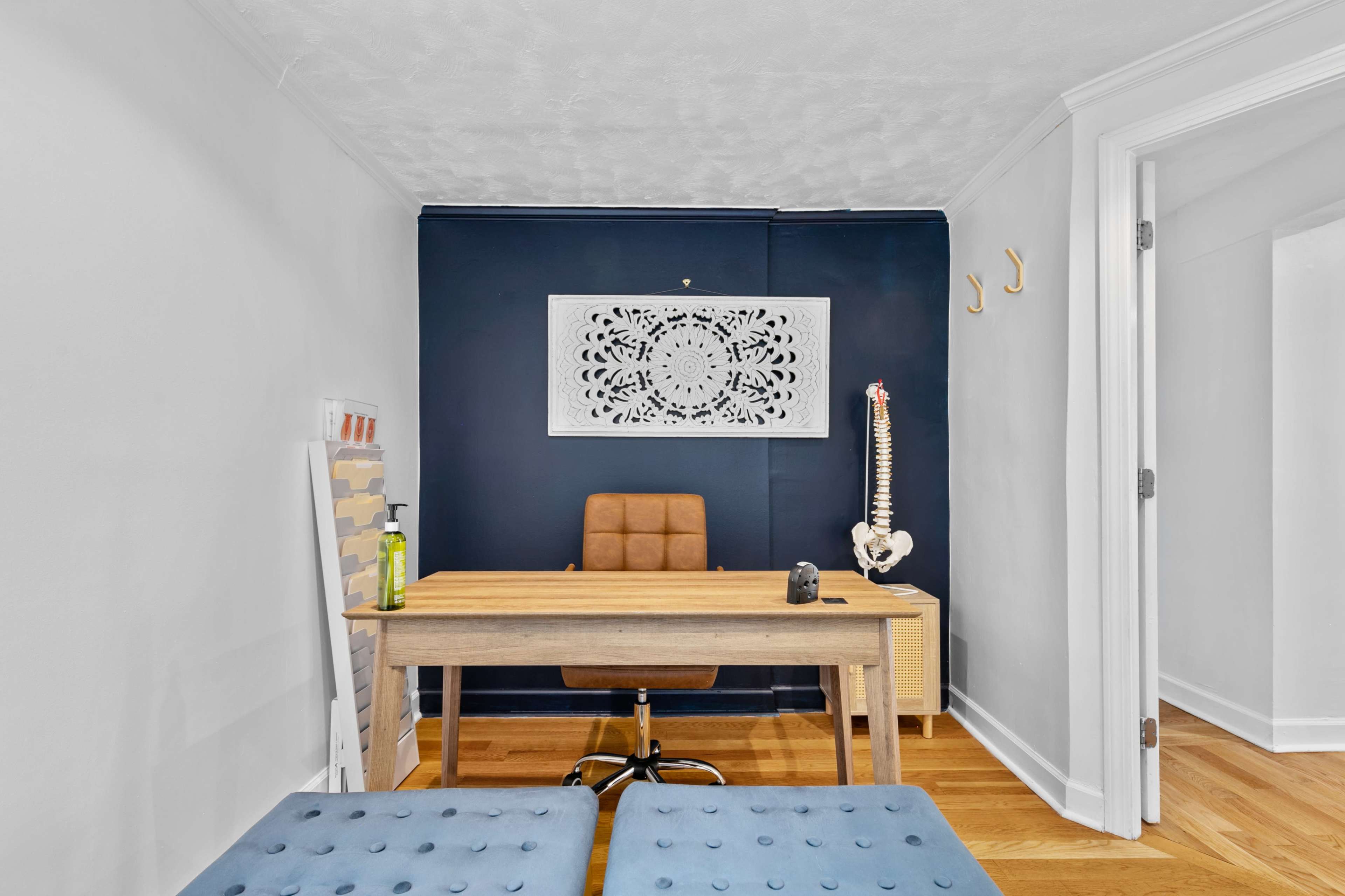 The image shows a small office space featuring a wooden desk with a chair, a decorative wall art piece, and a model of a human spine.