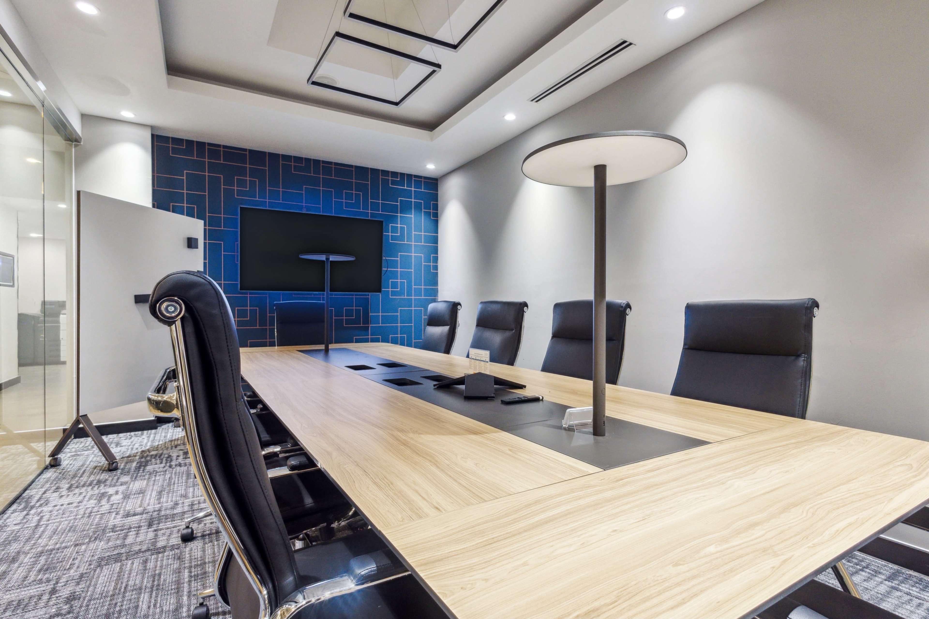 A modern conference room features a wooden table with black leather chairs, a wall-mounted screen, and a floor lamp against a blue patterned backdrop.