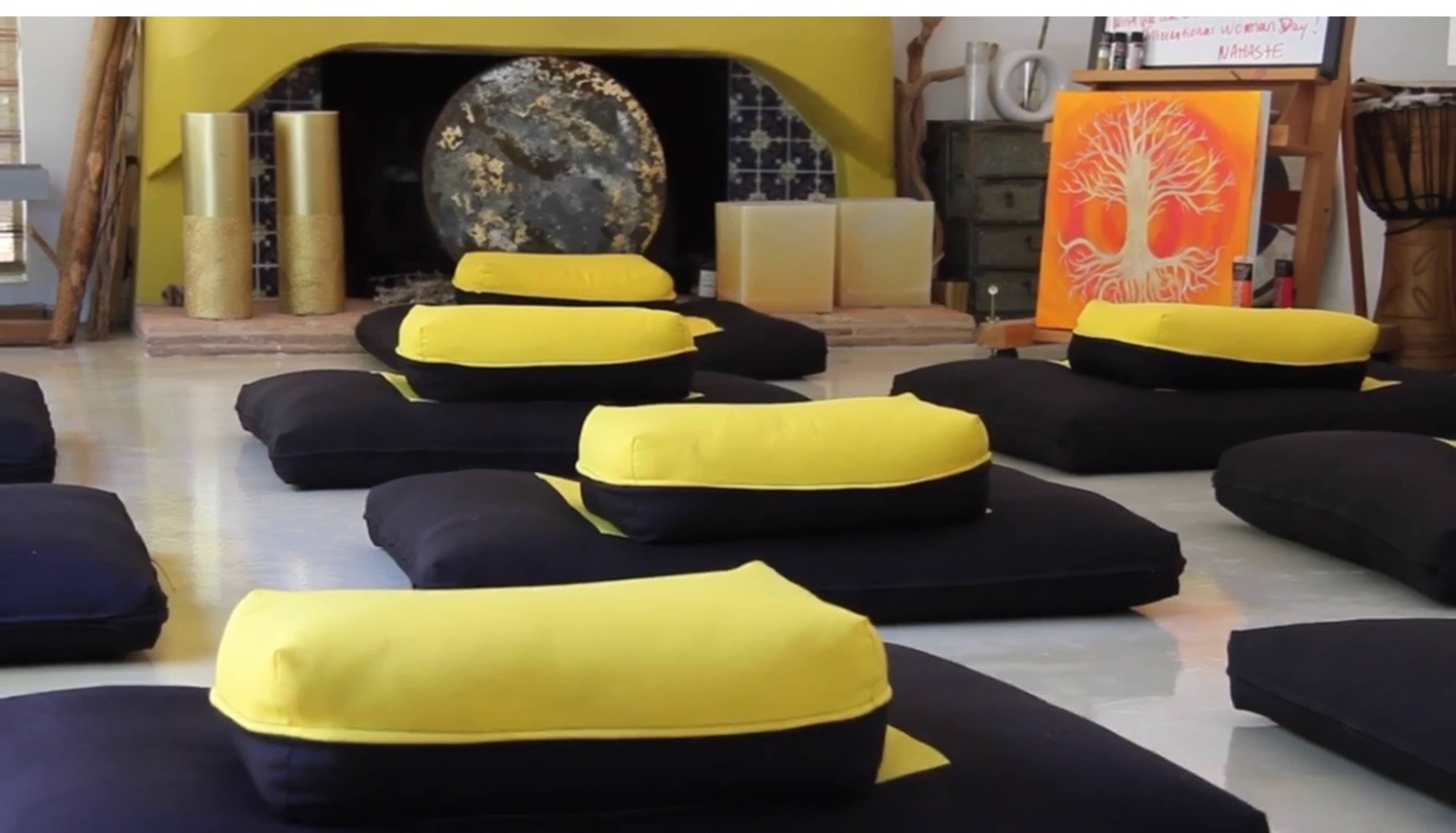 The scene shows a meditation space with several black cushions featuring yellow tops arranged on a polished floor, near a fireplace with decorative items and artwork on the walls.