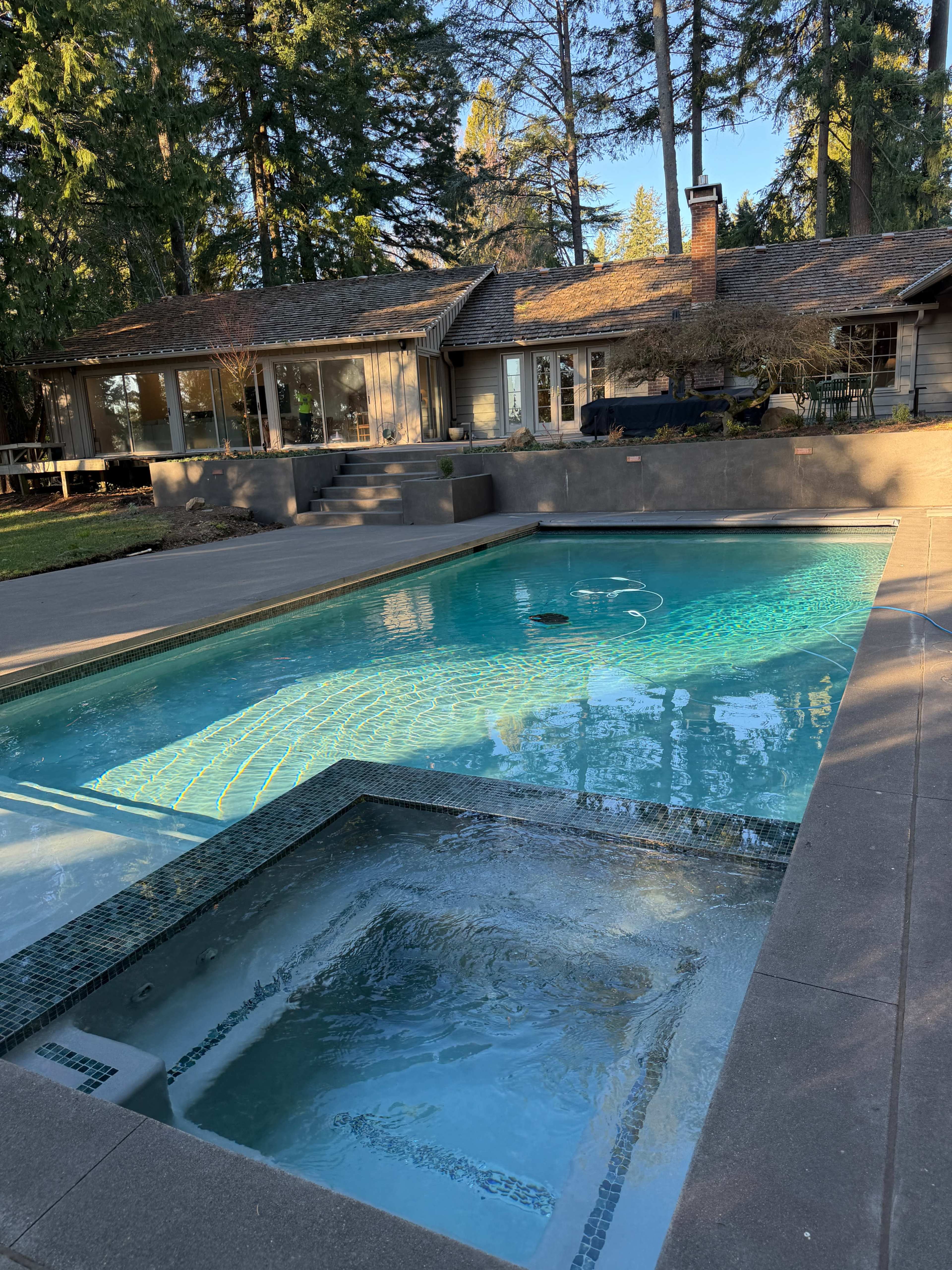 A swimming pool with an adjacent hot tub is situated in a backyard surrounded by tall trees and a house with large windows.