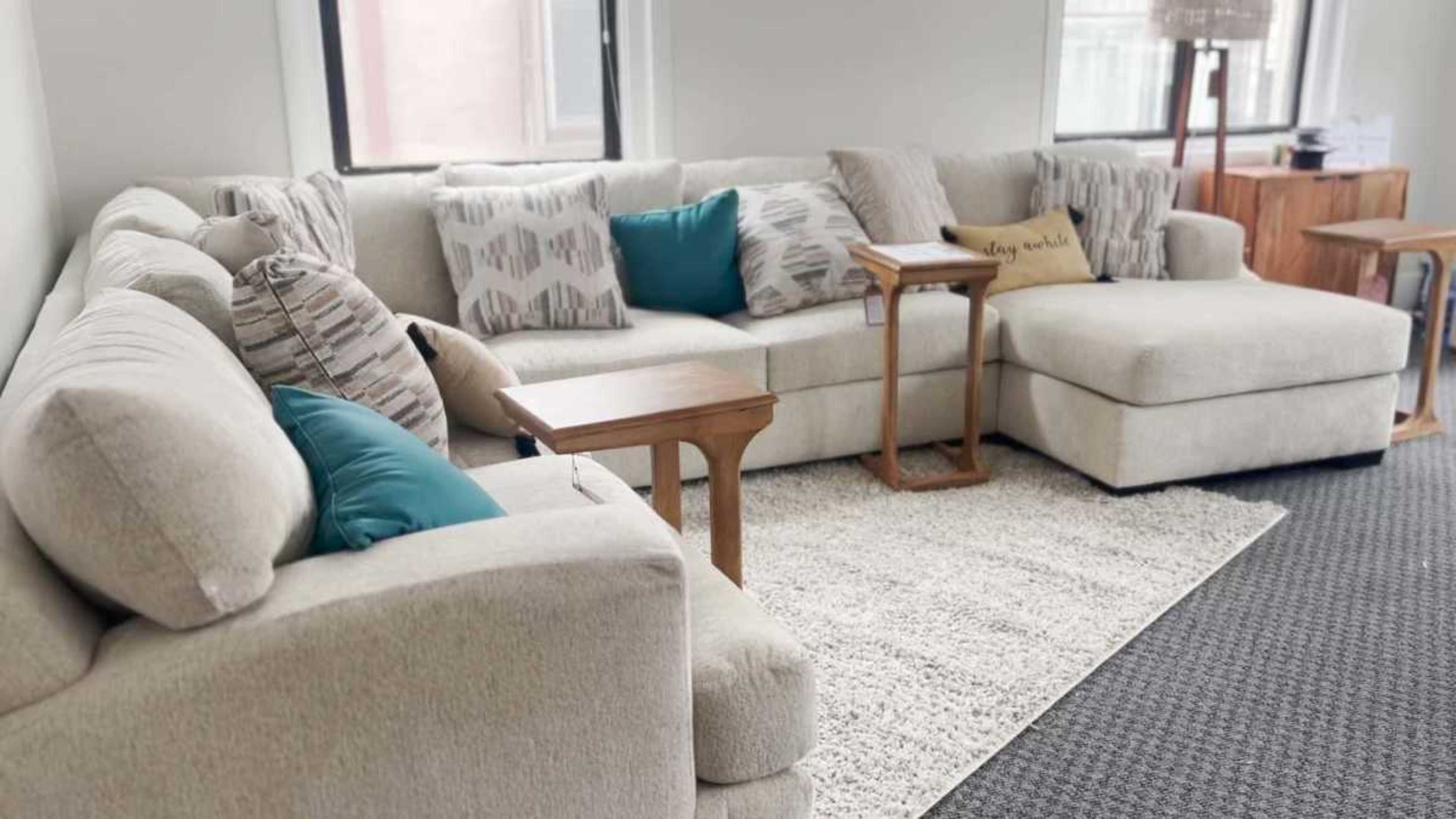 A spacious living room features a light-colored sectional sofa with patterned and solid-colored cushions, accompanied by two wooden side tables and a textured area rug.