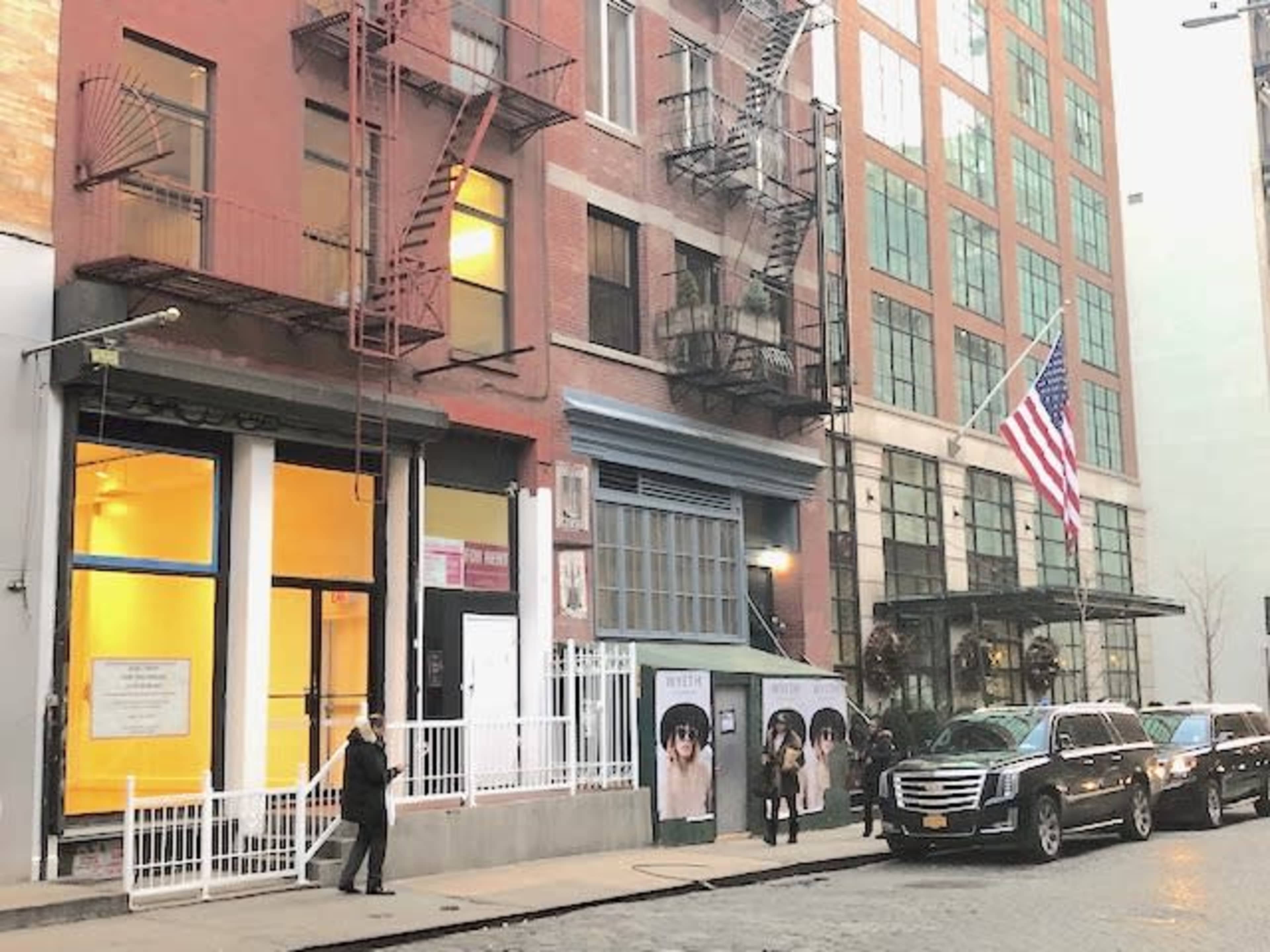 A brick building with fire escapes stands alongside a row of parked cars, with a large American flag displayed on the front.