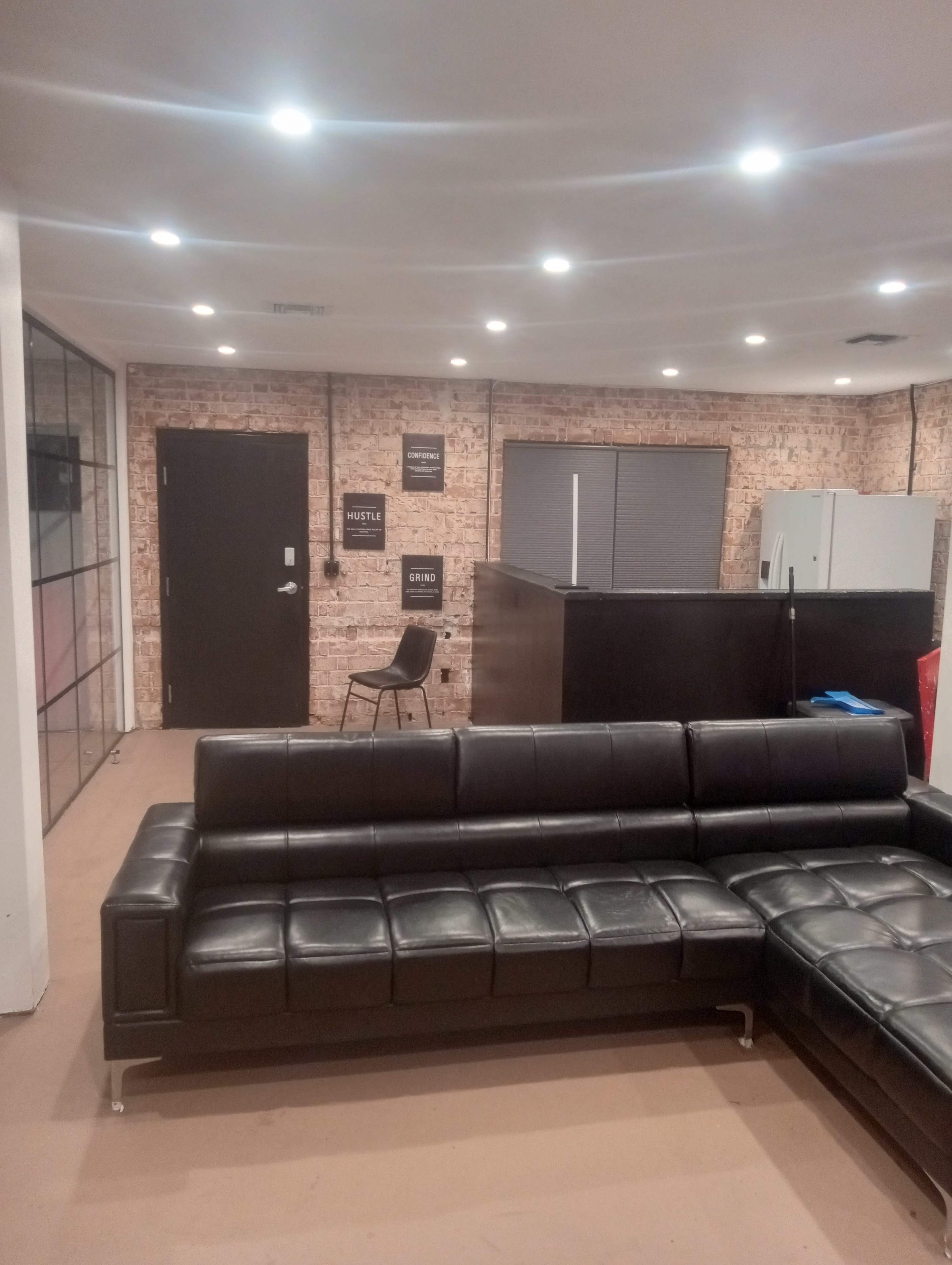 The image shows a modern office interior featuring a black sectional sofa, a glass partition, a door, and a kitchen area in the background.