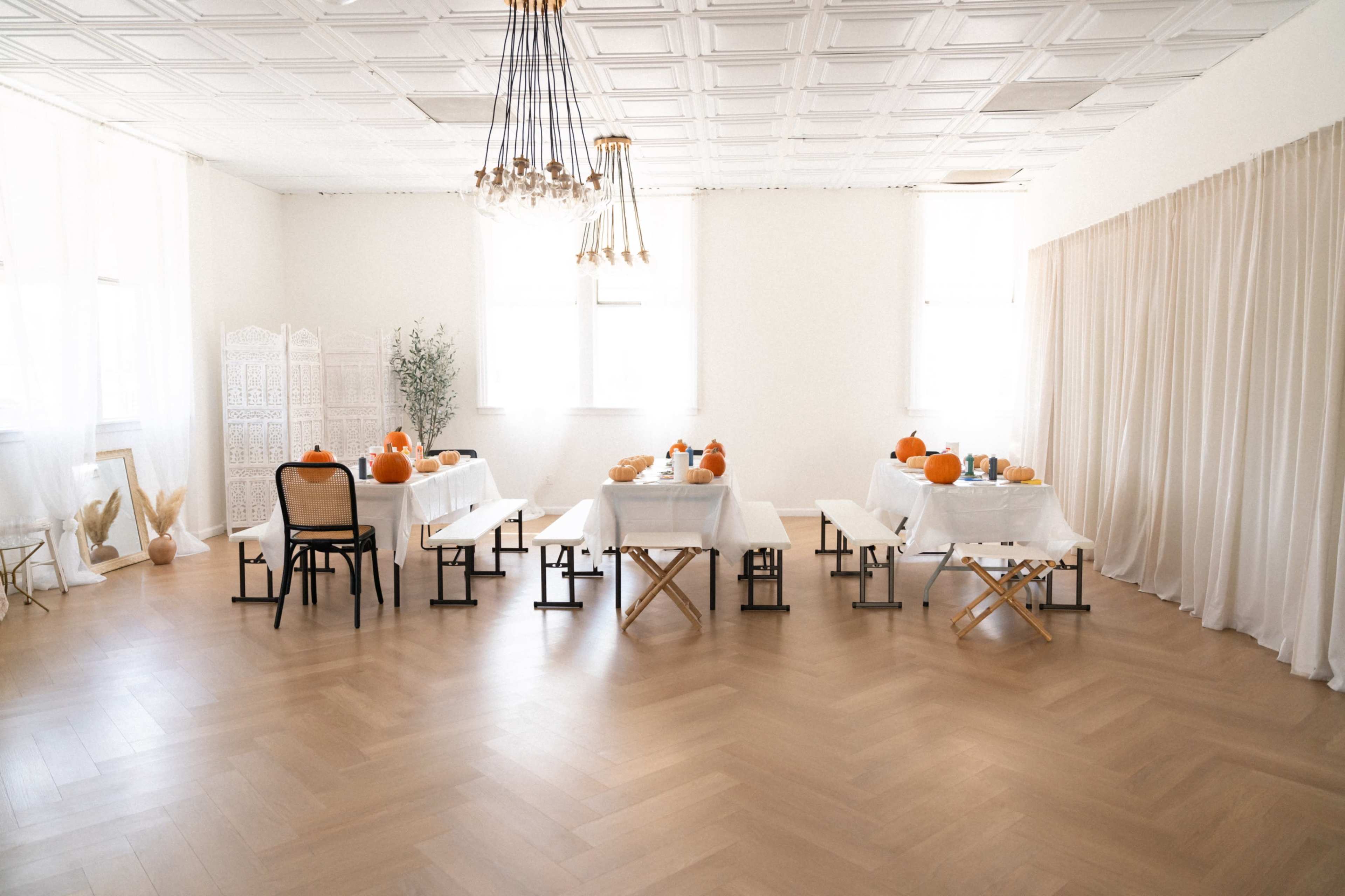 A bright, minimalist room set up for a gathering, featuring tables with white linens, pumpkins, and decorative elements, surrounded by chairs and a light wood floor.