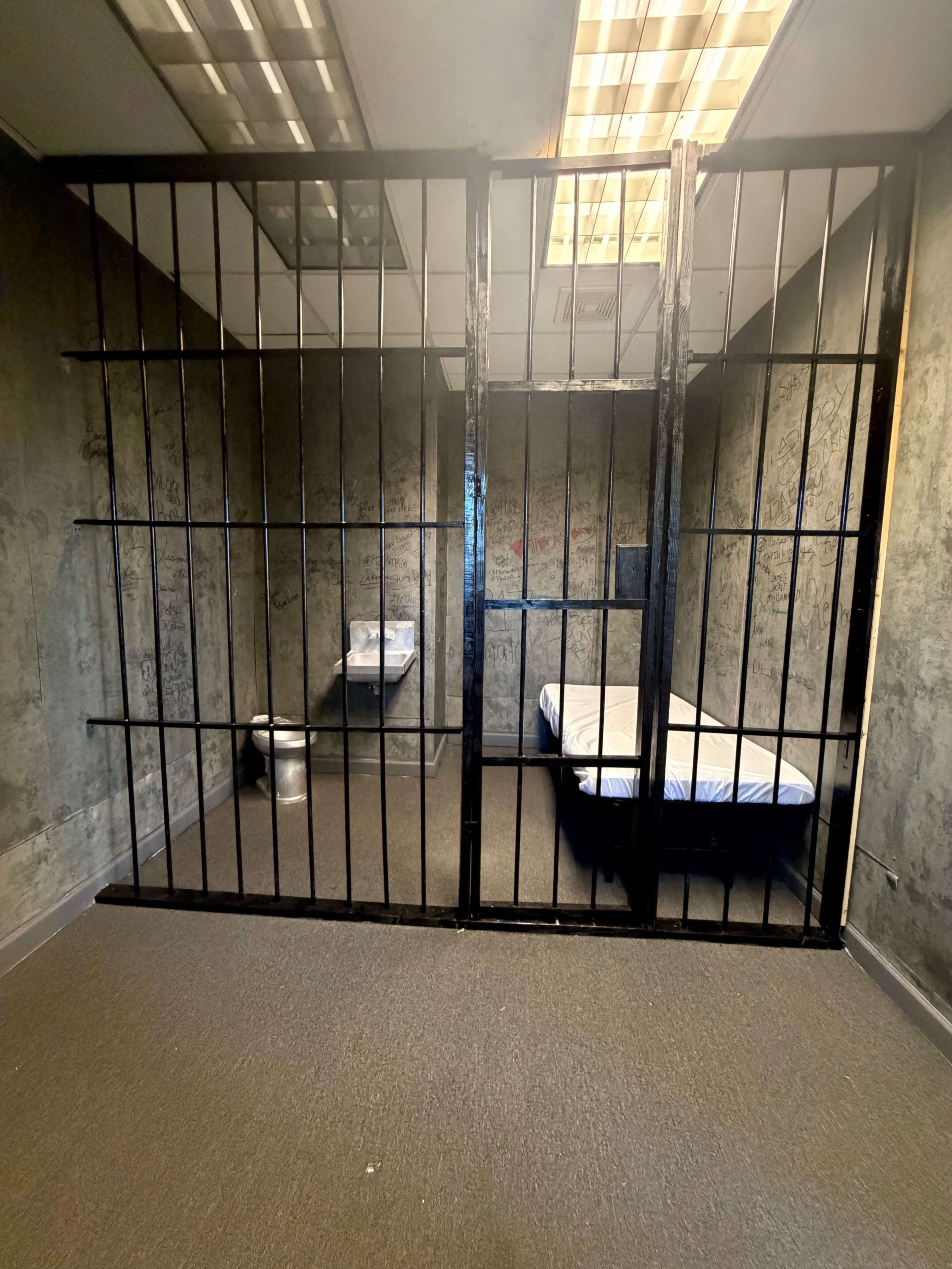 The image shows a small, sparse prison cell with barred partitions, a bed with a white mattress, and a toilet in the corner.