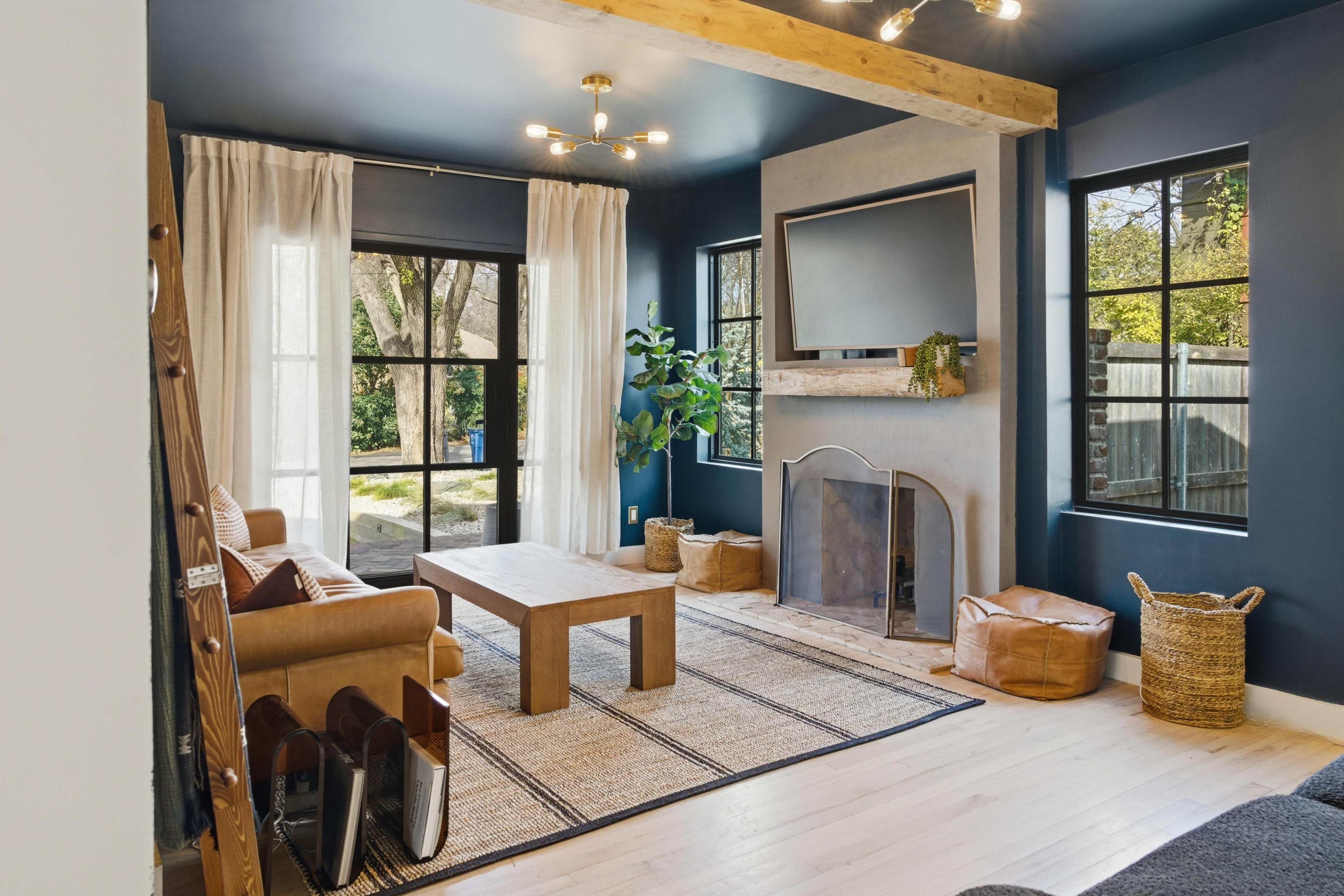 A modern living room features a leather sofa, a wooden coffee table, and large windows, with decorative plants and a flat-screen TV mounted on a concrete wall.
