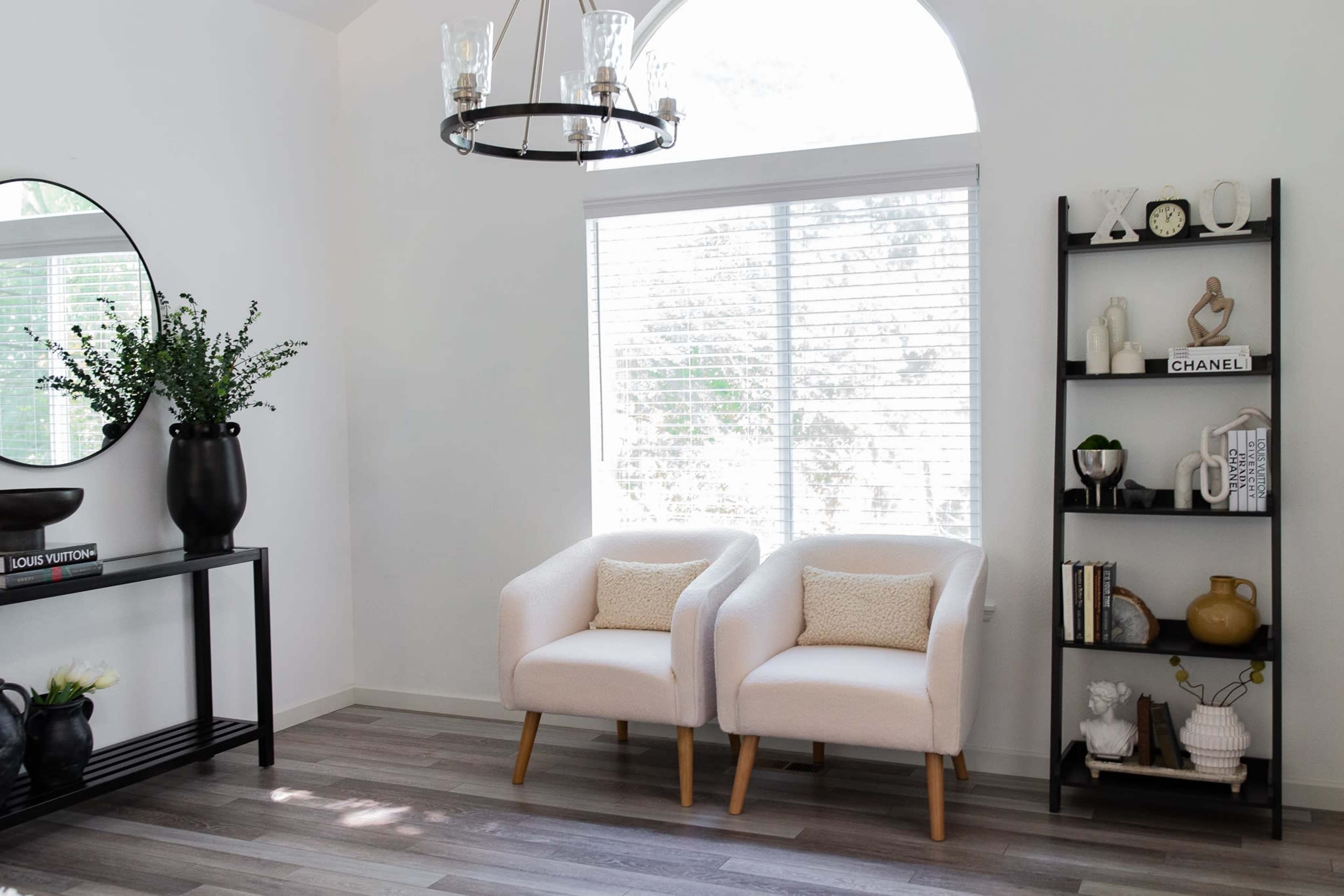 A well-lit living room features two white armchairs next to a black shelving unit and a round mirror above a black console table.