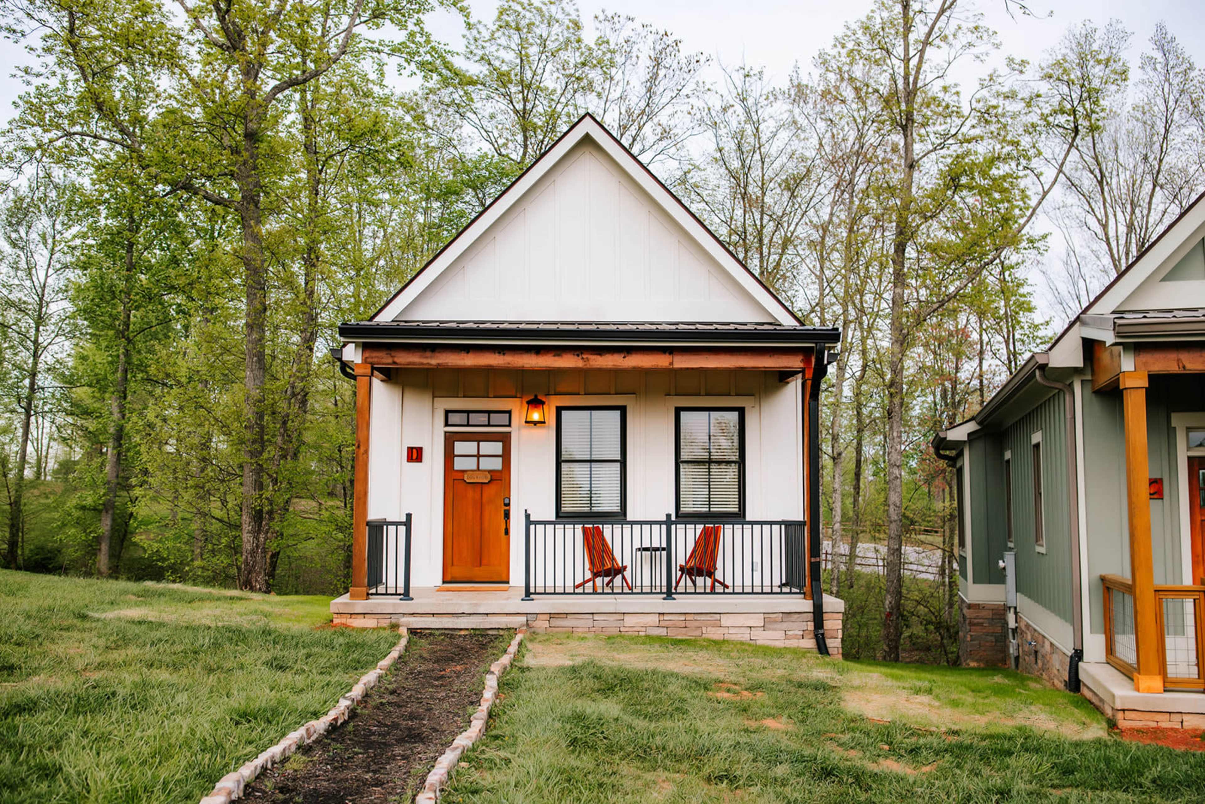 A small, modern house with a white exterior and a wooden front door is set among trees, featuring a porch with two red chairs.