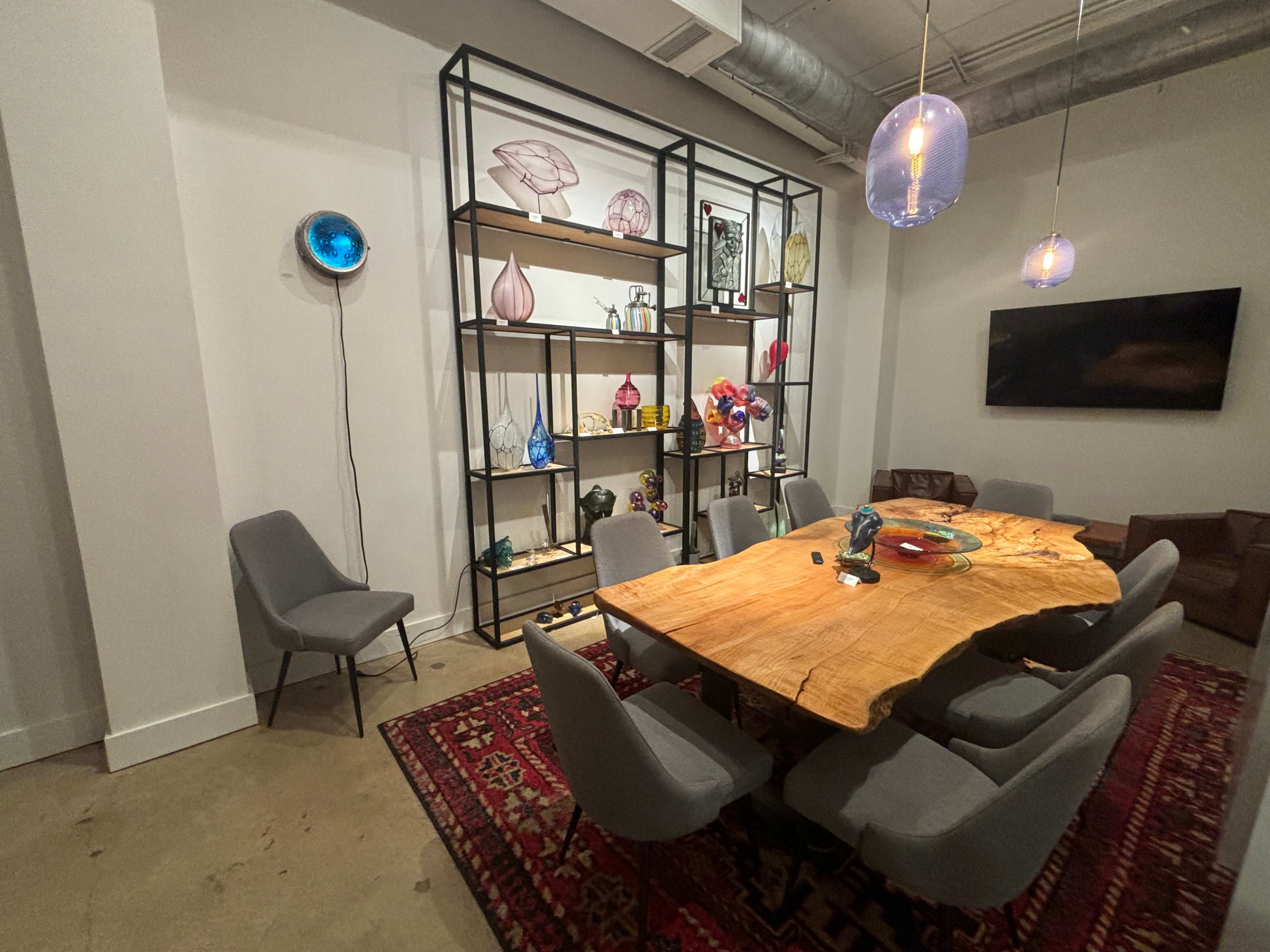 The image shows a modern dining area featuring a large wooden table surrounded by gray chairs, with a decorative shelving unit displaying various colorful items in the background.