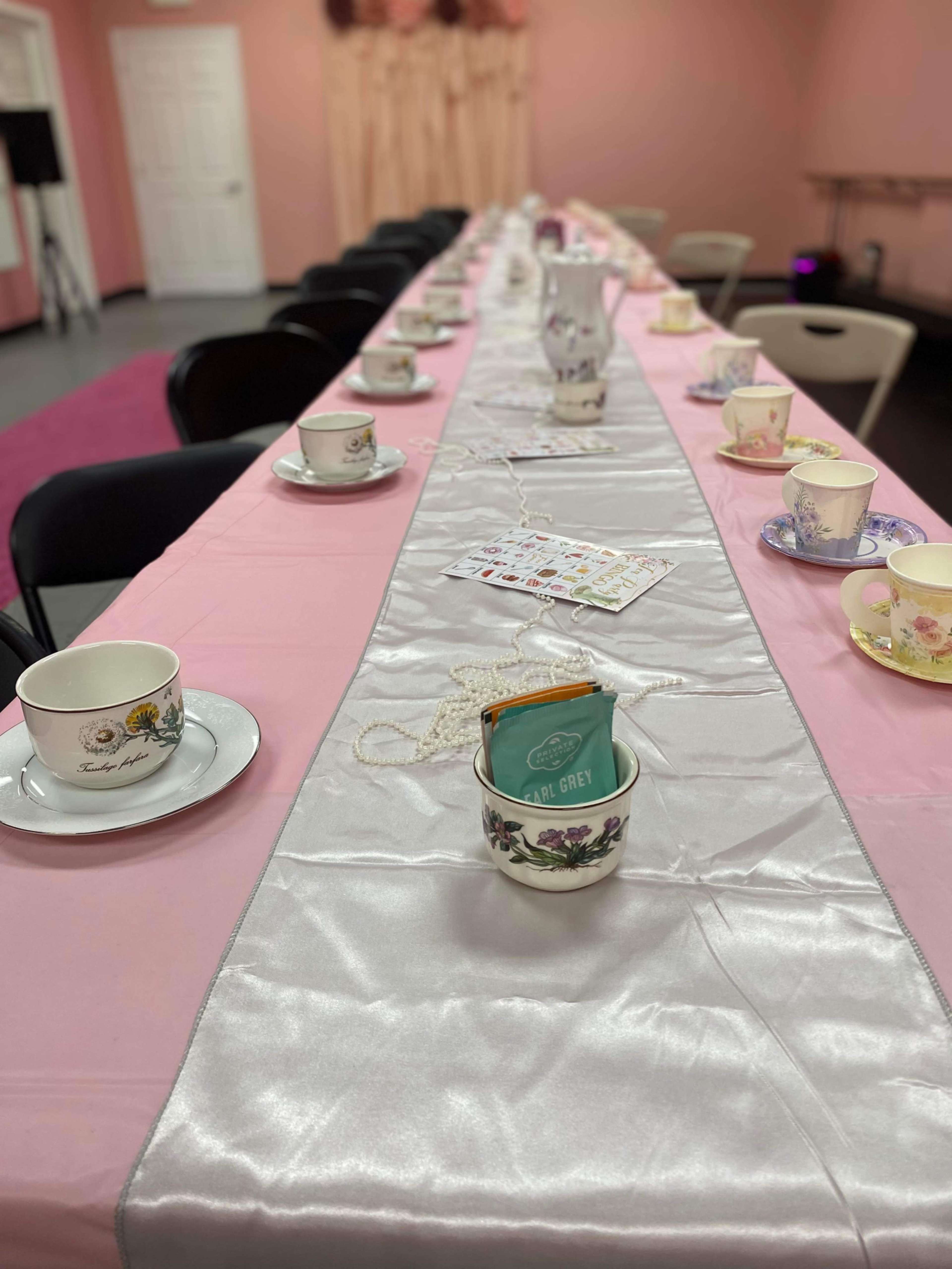 A long table is set with a pink tablecloth, adorned with vintage teacups and snacks, against a backdrop of pale pink walls.
