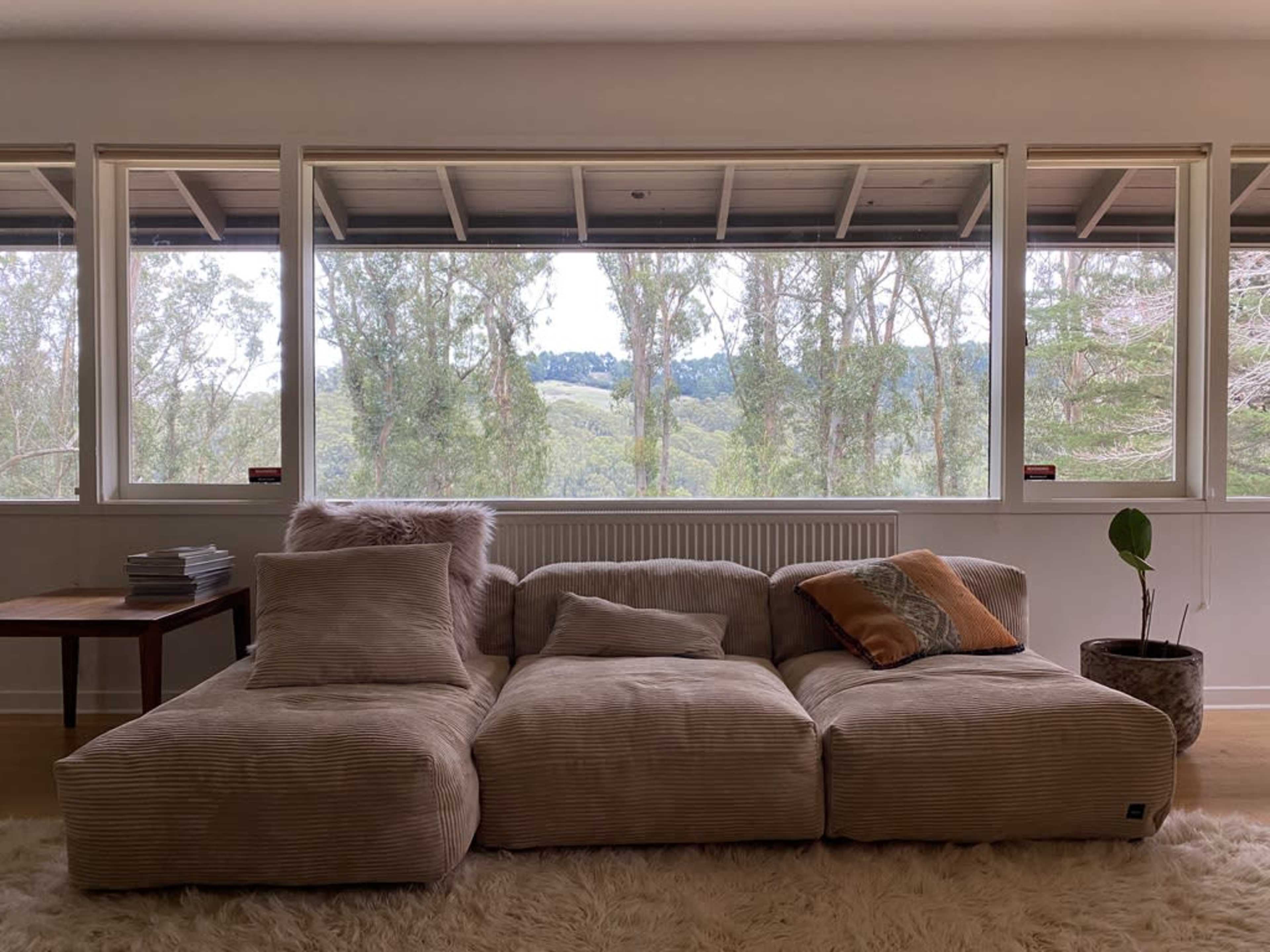 A large, upholstered sectional sofa is positioned in front of wide windows that offer a view of lush green hills and trees.