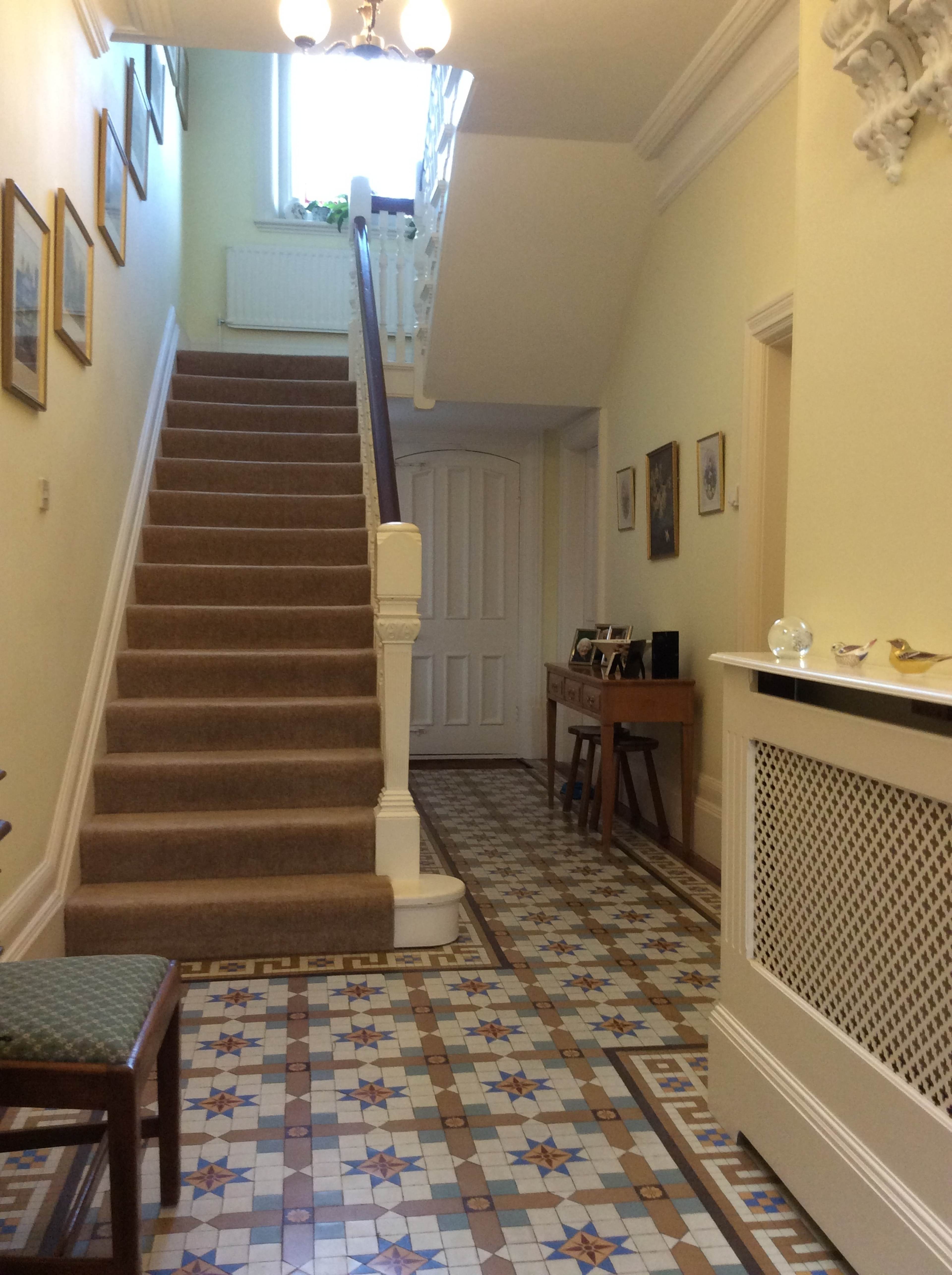 A hallway with a staircase leading to an upper floor, framed by wall-mounted pictures and a patterned tile floor.