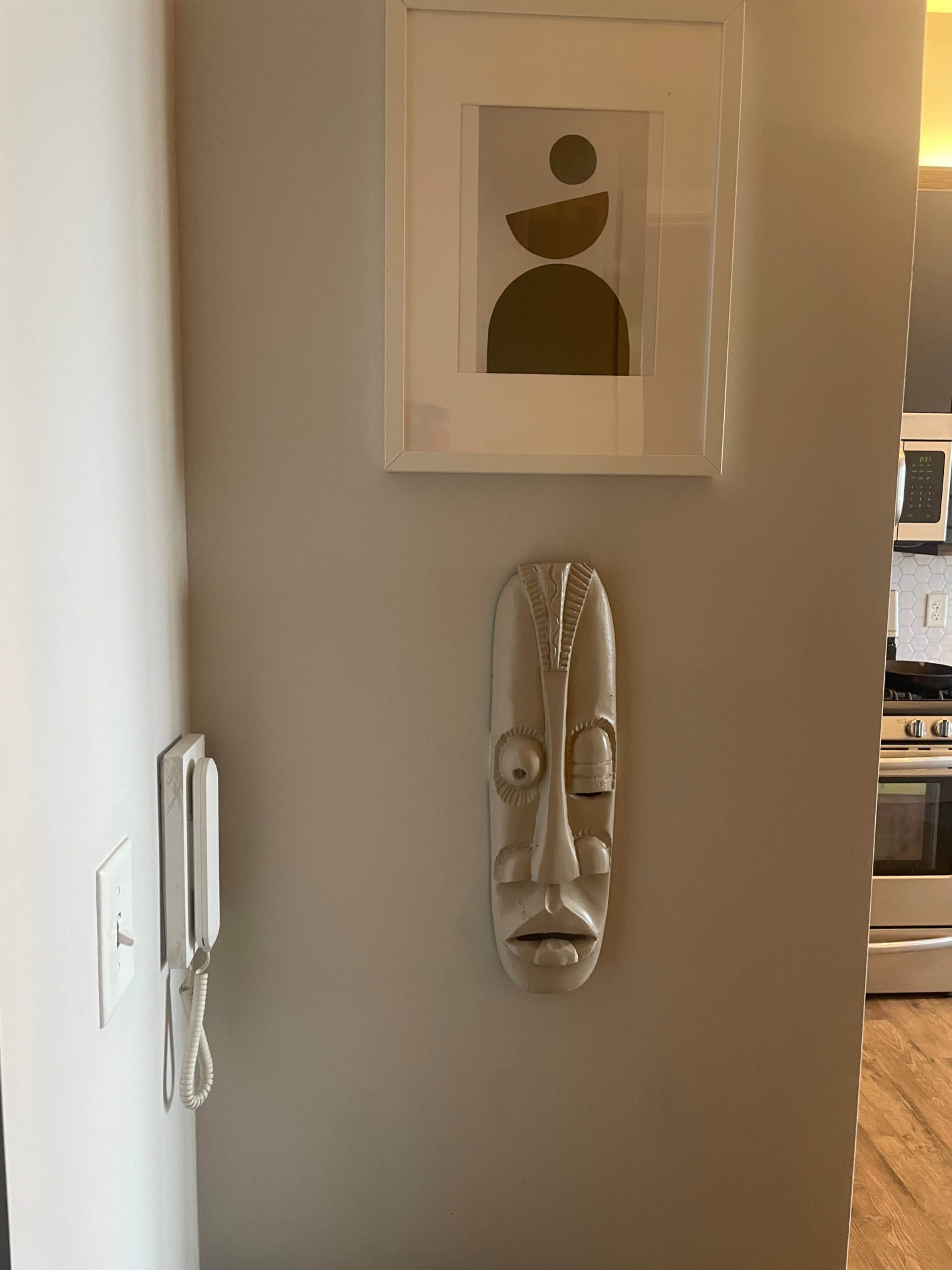 A white wall features a framed abstract artwork above a carved white mask, next to a wall-mounted telephone.
