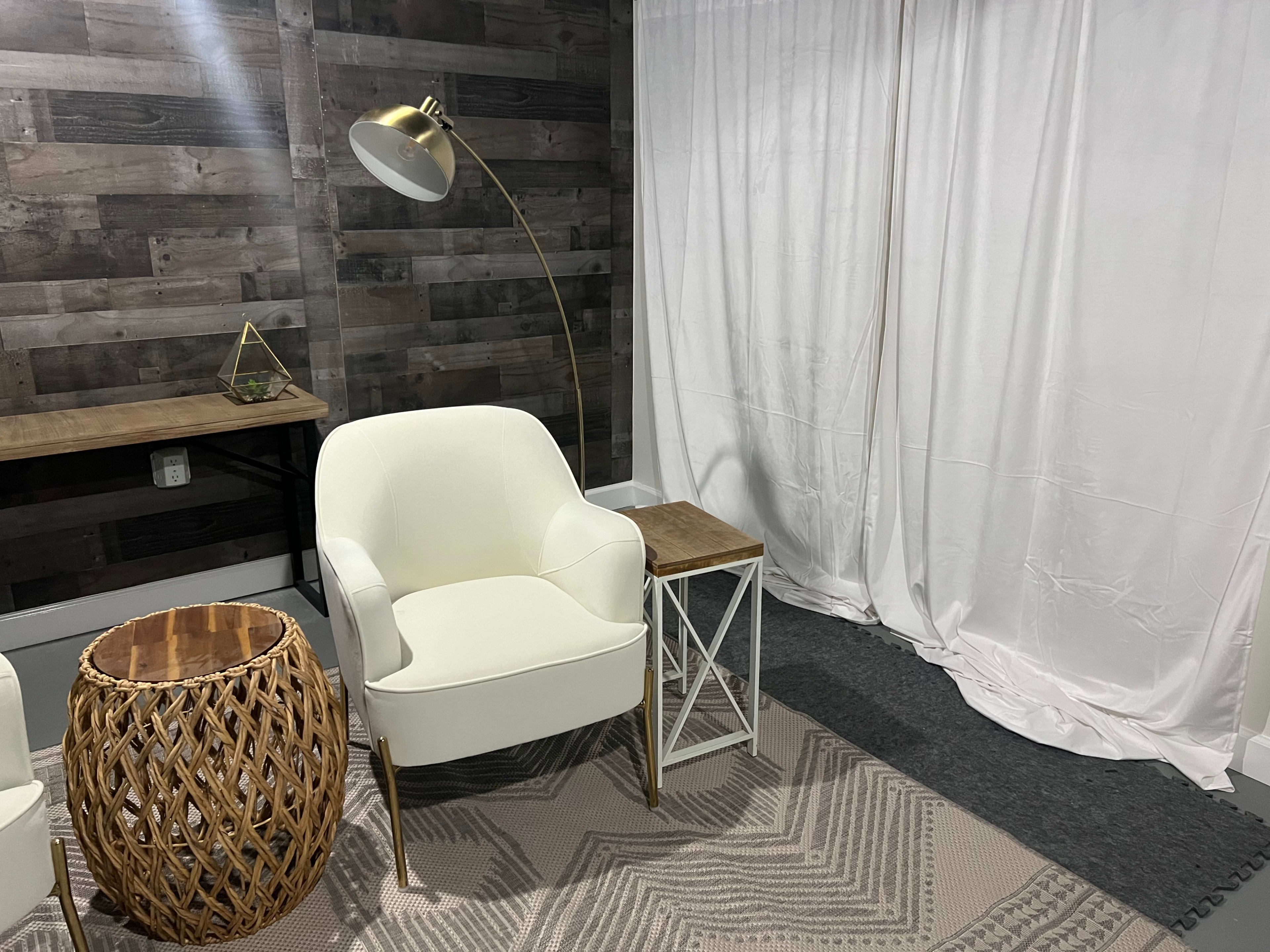 The scene features a cozy seating area with a white armchair, a wooden side table, a woven basket, and a floor lamp against a rustic wooden wall and a white curtain.